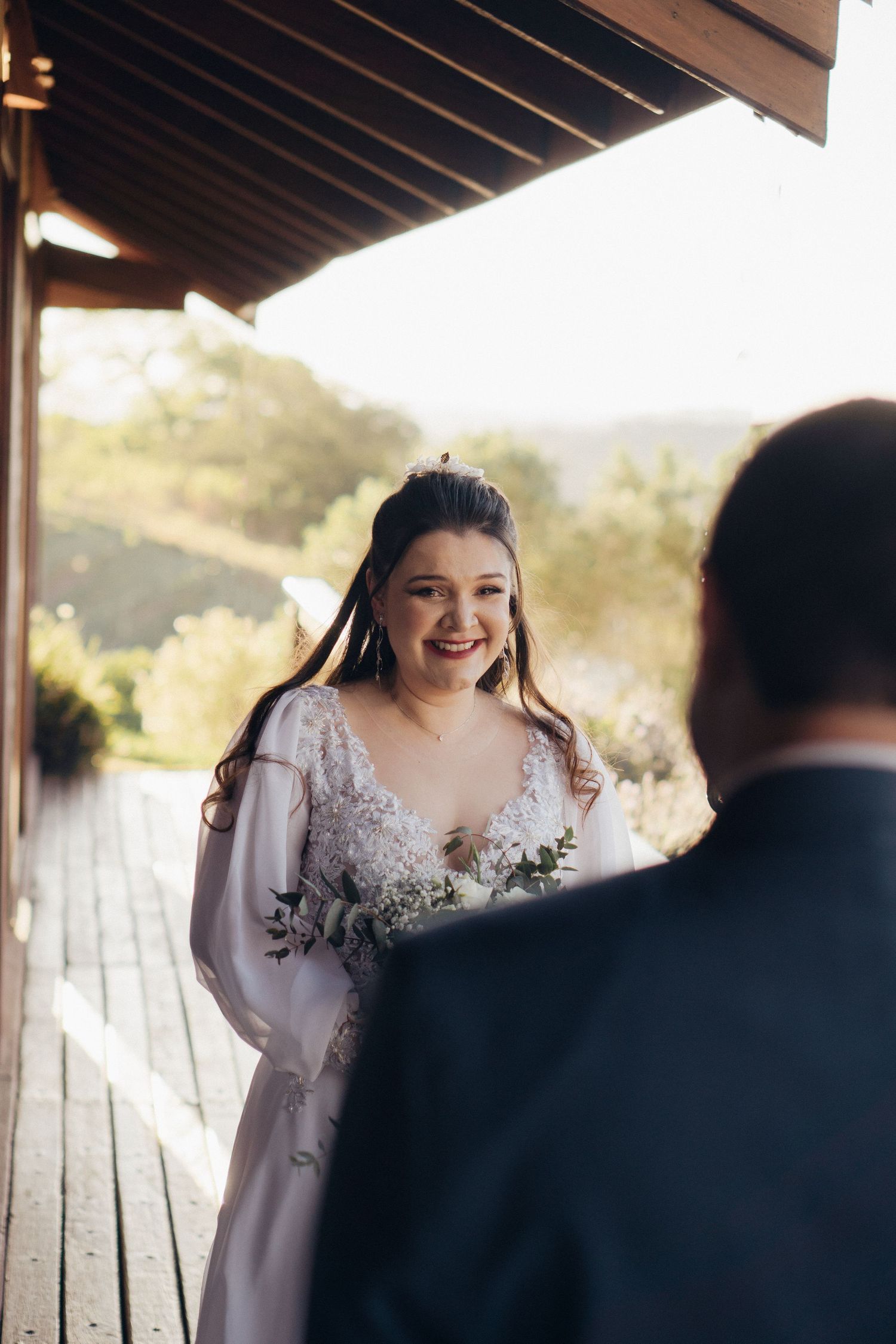 A bride in a white lace dress stands on a wooden deck during golden hour.