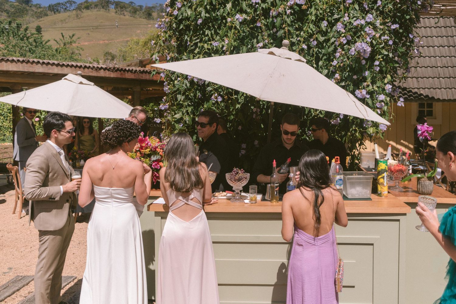 Convidados se reúnem ao redor de um bar ao ar livre sob guarda-sóis brancos, em casamento ao ar livre em cenário montanhoso