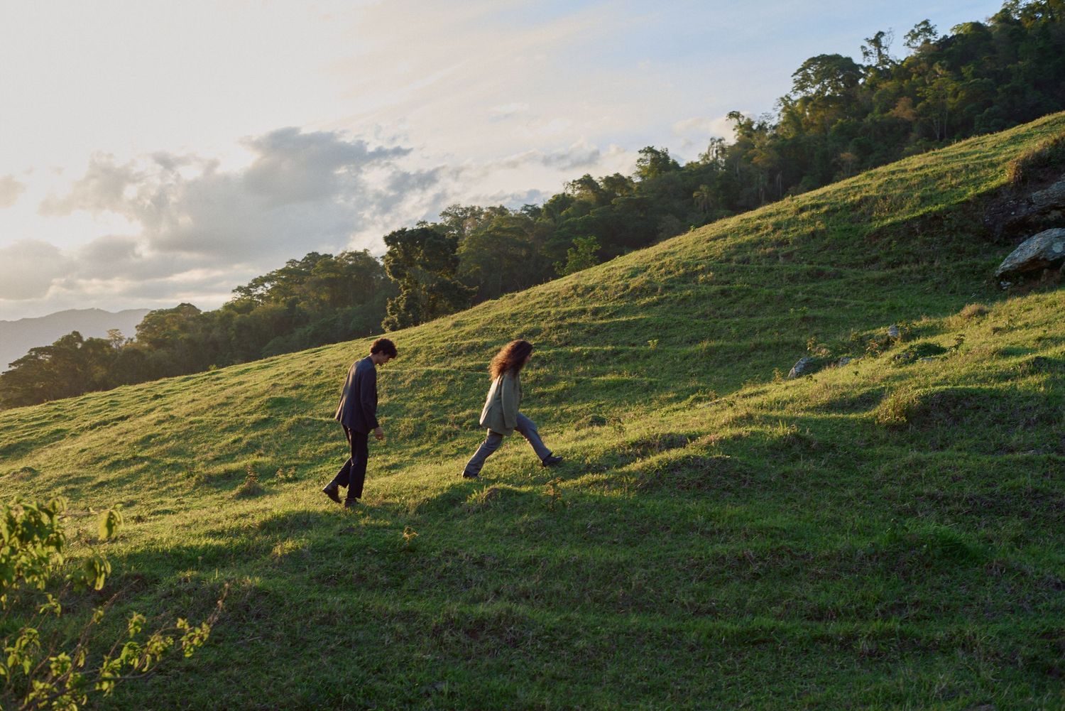 Duas pessoas caminham por uma encosta gramada ao pôr do sol, projetando longas sombras sobre a superfície verdejante.