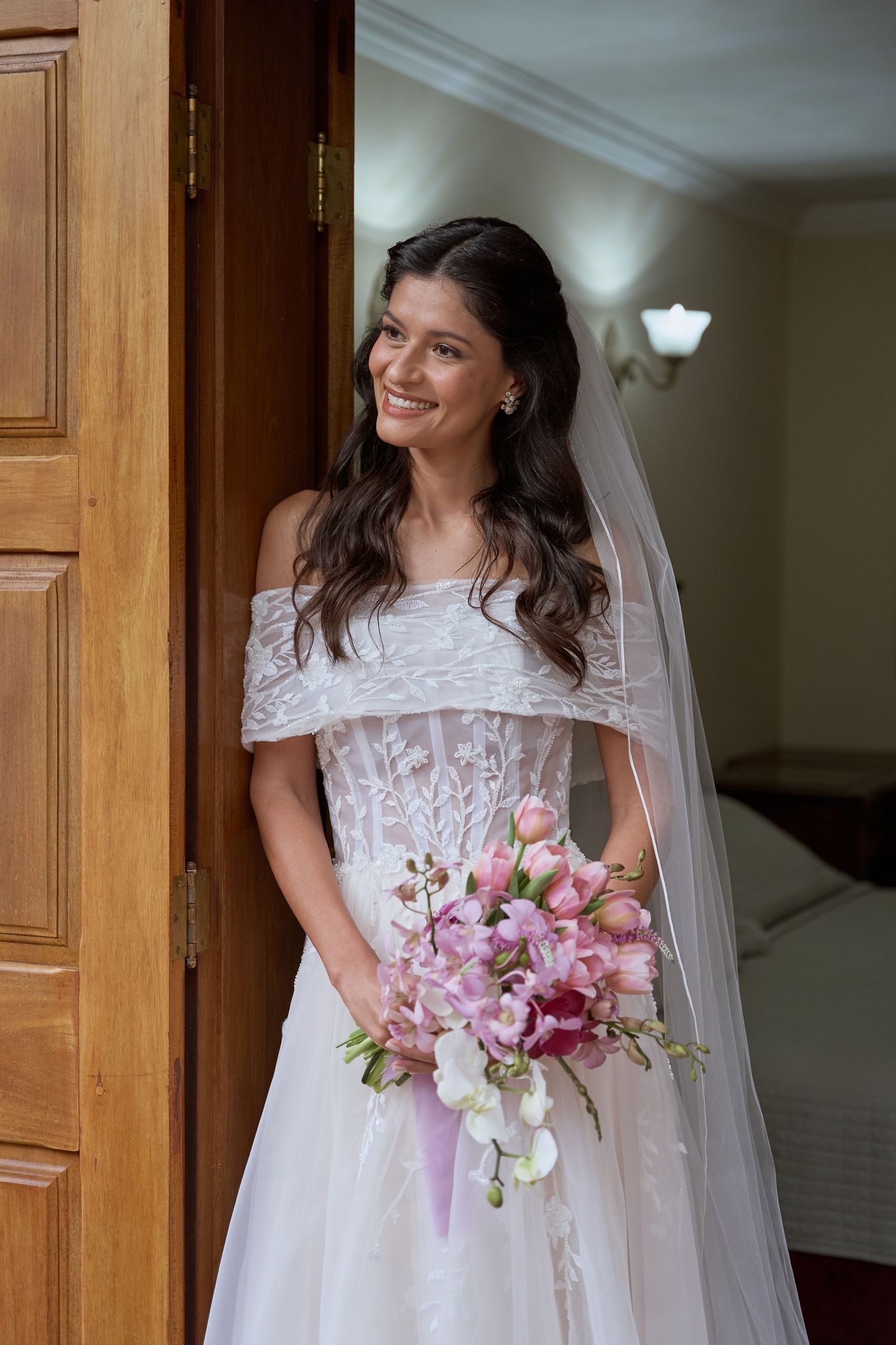 Uma noiva com um vestido de renda branca segura um buquê rosa e branco enquanto está em uma porta.