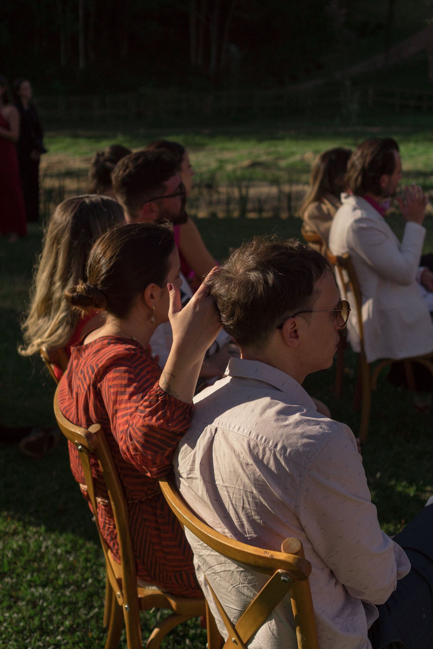 Detalhe de convidados relaxados durante a cerimônia de casamento ao ar livre, fotografado de forma natural e sem interferência, ao pôr do so
