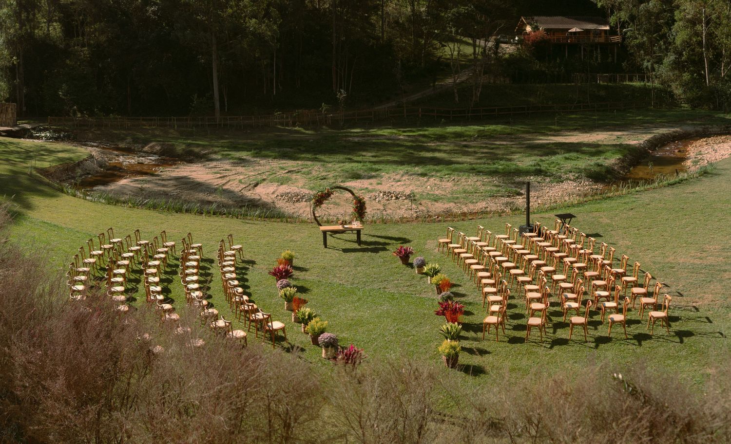 Fileiras de cadeiras de madeira em estilo de anfiteatro em colina gramada, com arco de madeira ao centro, em casamento ao ar livre
