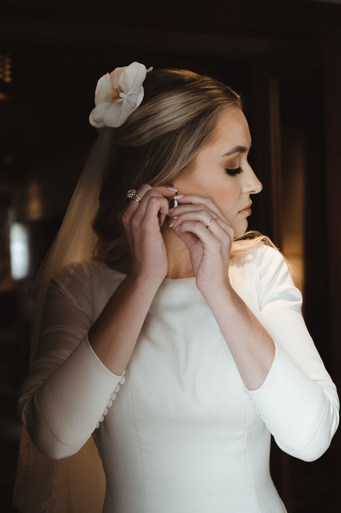 Uma noiva com um vestido de casamento branco de mangas compridas ajusta seus brincos em uma sequência de fotos elegantes dos preparativos.