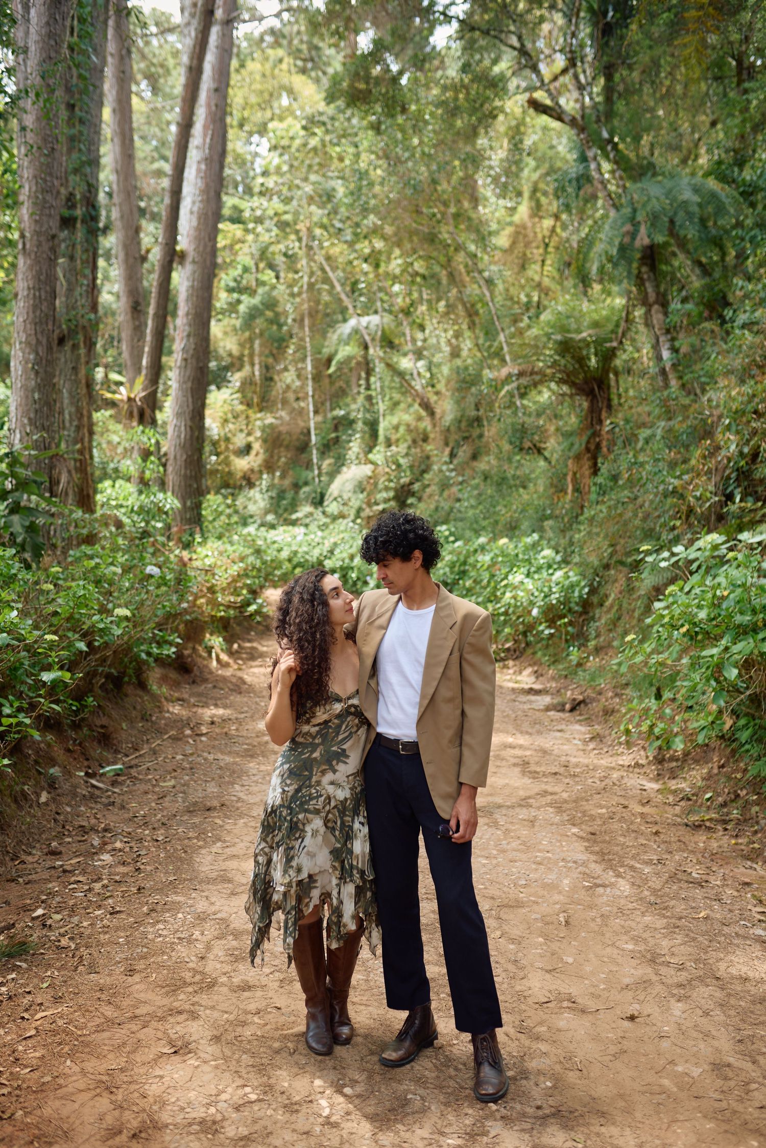 Um casal caminha junto por uma trilha de terra em meio a uma floresta exuberante durante uma sessão de fotos romântica ao ar livre.