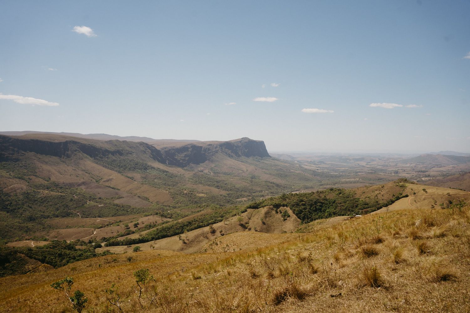 Uma vista panorâmica de colinas onduladas e montanhas marrons se estende por uma vasta paisagem desértica sob um céu azul. Serra da Canastra