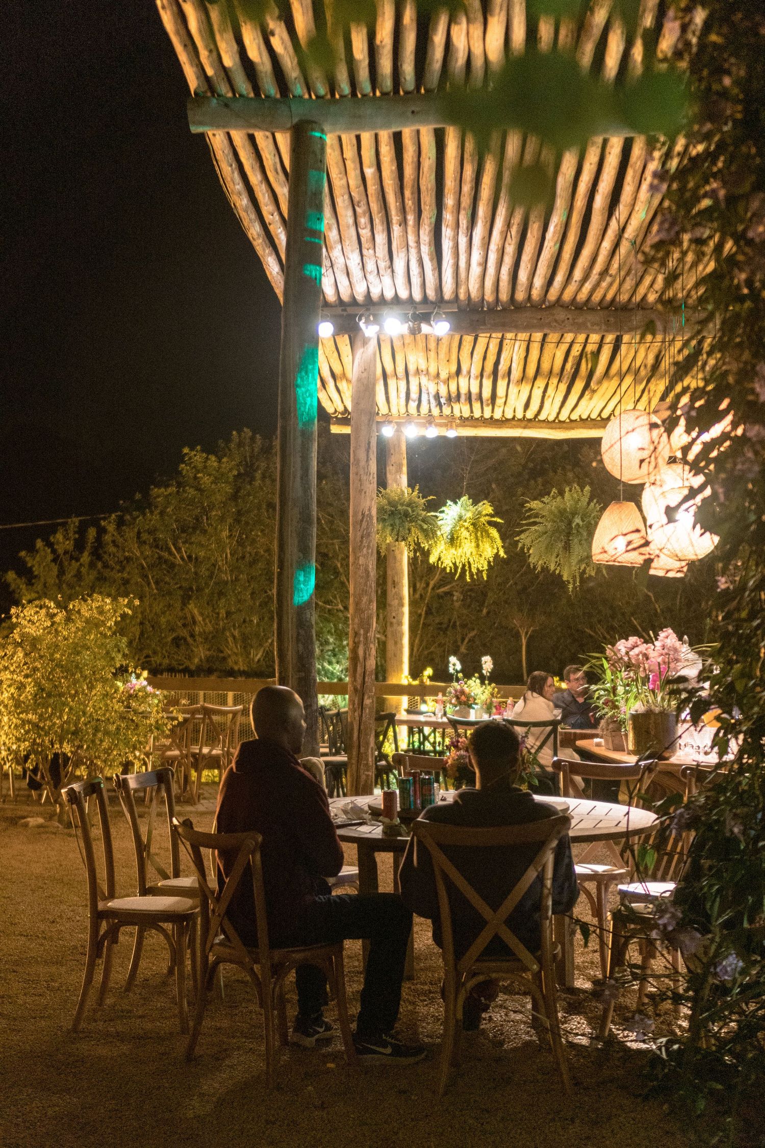 Festa de casamento ao ar livre à noite, com pérgola rústica de madeira, luzes pendentes e convidados jantando sob iluminação aconchegante