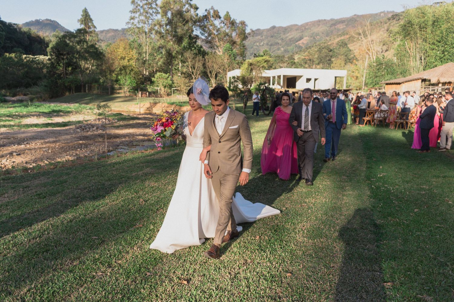 Casal radiante caminhando pelo gramado, em uma cerimônia de casamento ao ar livre, com montanhas ao fundo e convidados festivos