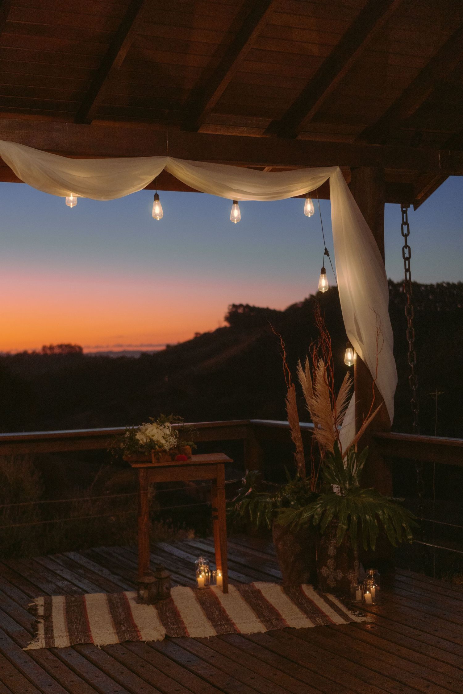 Deck rústico decorado com luzes de corda e cortinas brancas ao pôr do sol.
