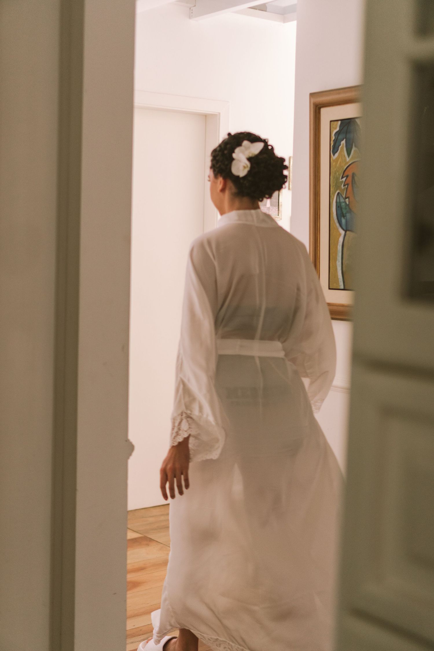 Noiva em roupão branco e acessório floral no cabelo, em pé na porta de um corredor iluminado de interior