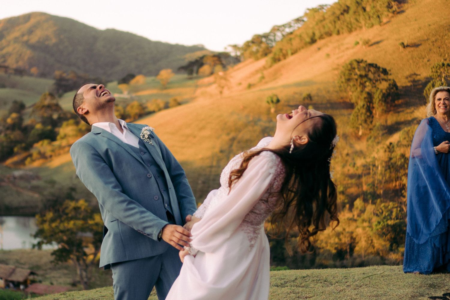 Um casal dança alegremente em um cenário de montanha durante a hora dourada, com vestidos esvoaçantes e trajes formais.