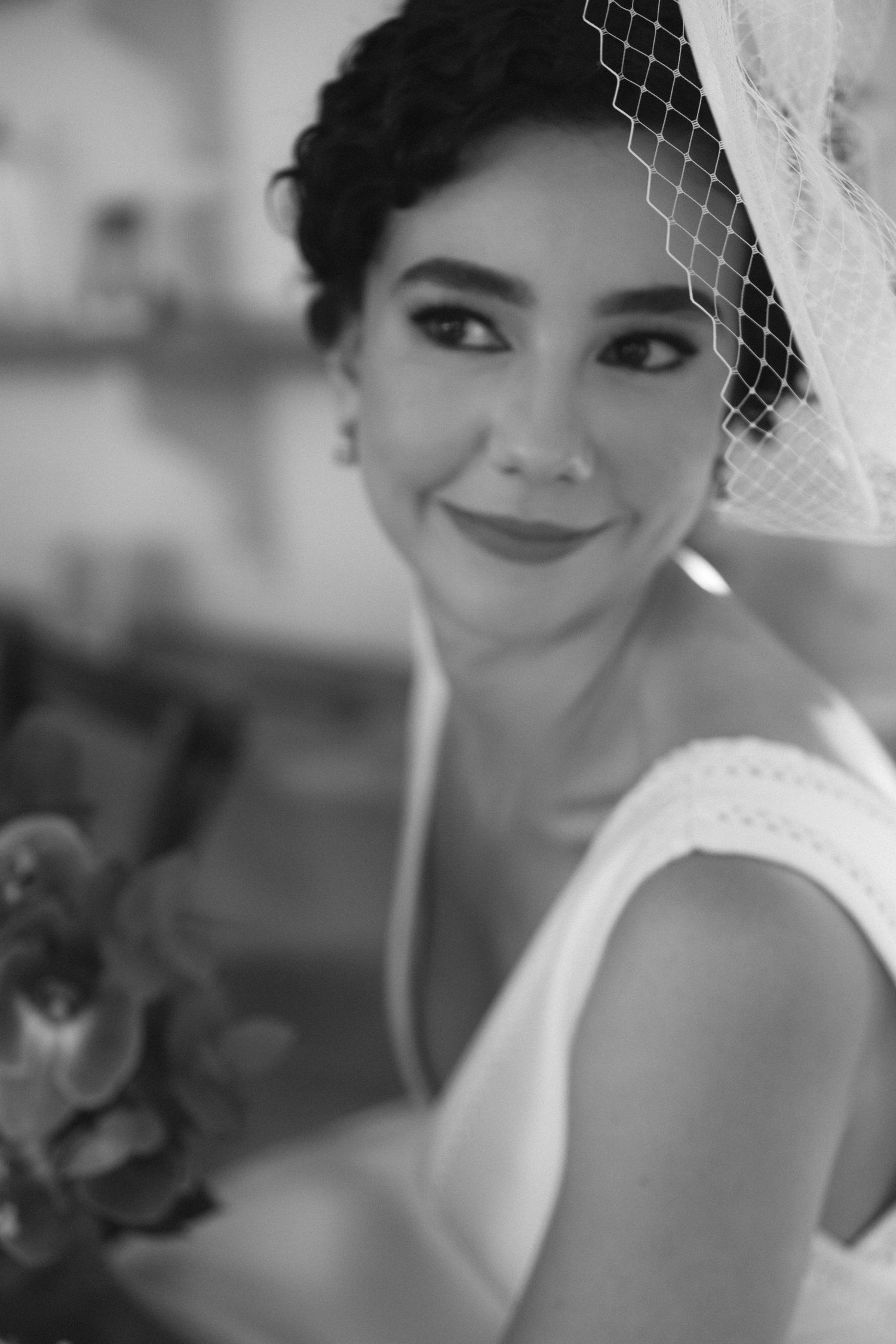 Retrato em preto e branco de noiva com véu estilo birdcage e vestido branco, sorrindo calorosamente para a câmera