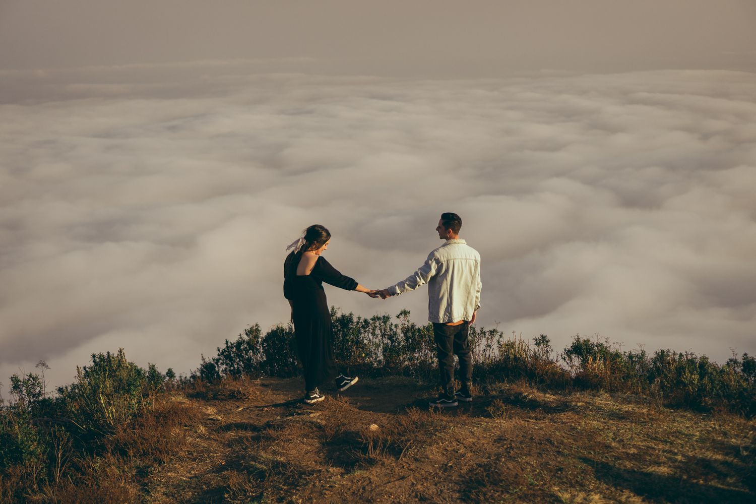 Duas pessoas de mãos dadas no topo de uma montanha, acima das nuvens, ao pôr do sol, em meio a uma vista panorâmica deslumbrante.