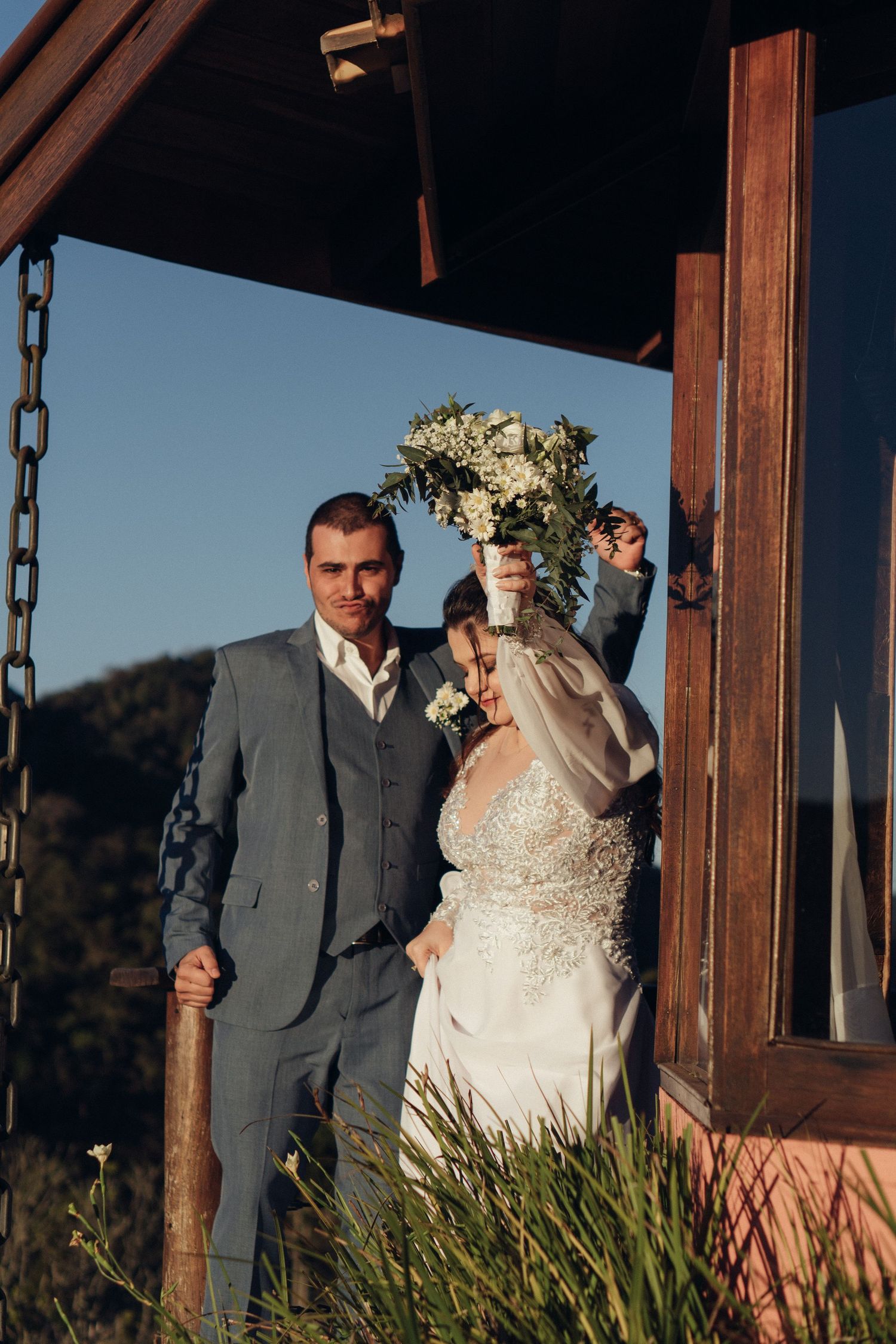 Um casal posa em uma varanda coberta com um buquê de noiva em um cenário montanhoso.