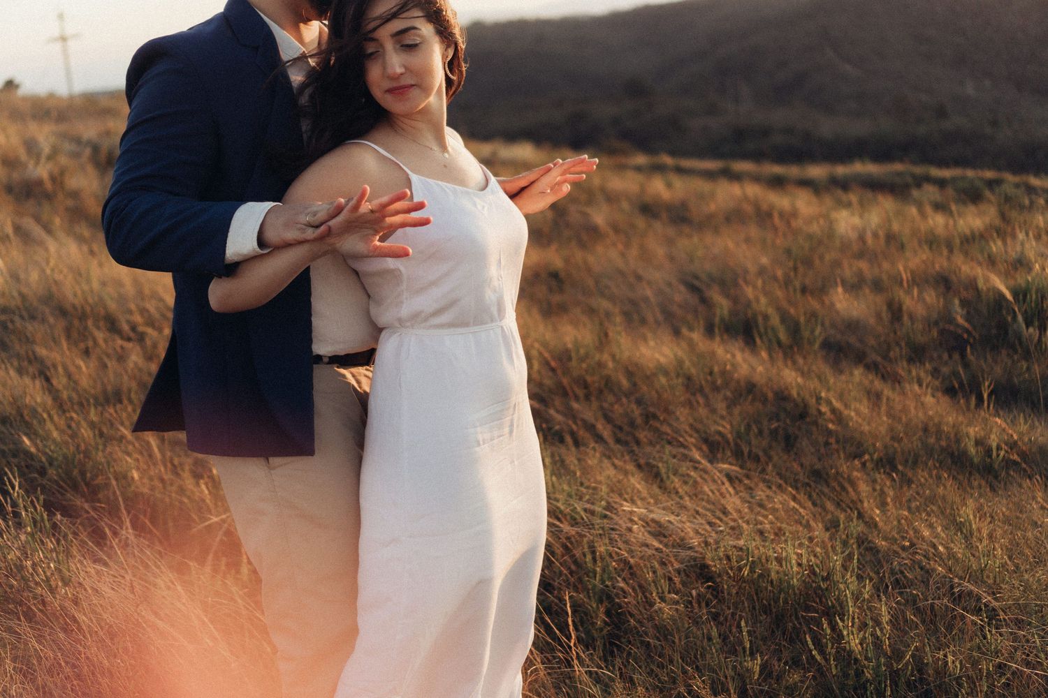 A couple embraces in a golden field at sunset, with the person in white showing a visible baby bump.