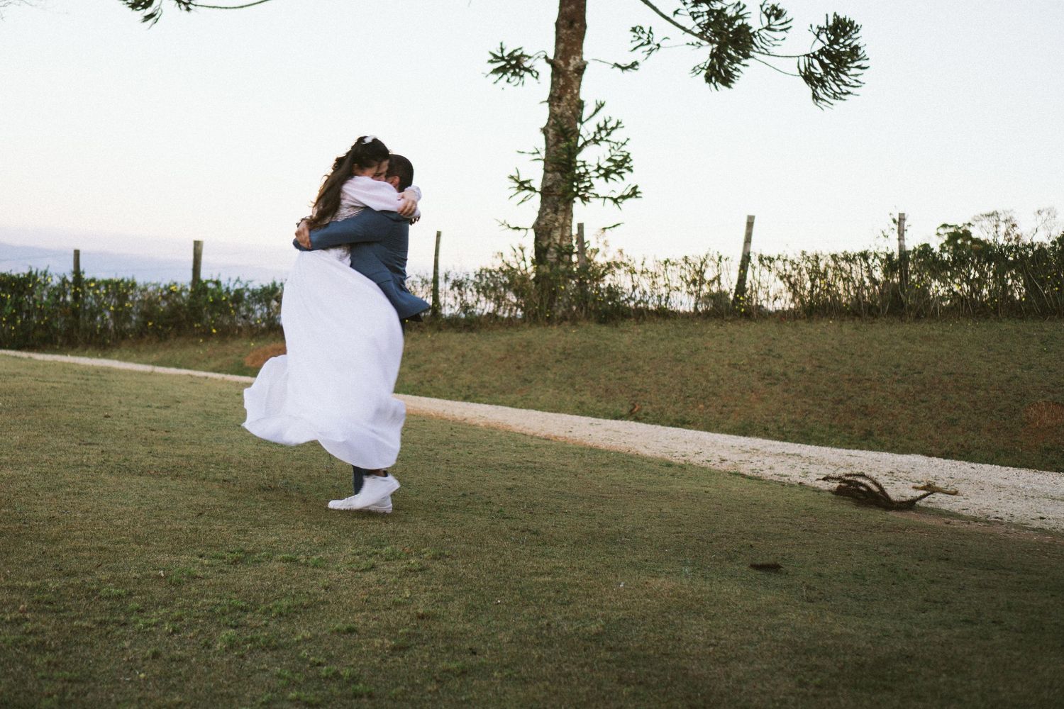 Um casal se abraça e gira em uma trilha gramada no topo de uma colina com vistas panorâmicas das montanhas ao fundo.