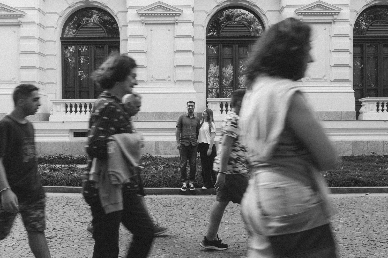 Casal olha pra câmera enquanto pessoas passam em primeiro plano, borradas © Clara e Matheus Fotógrafos de Casamentos