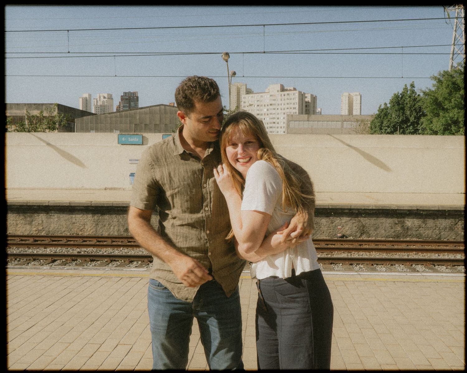 Casal se abraça e olha pra câmera com trilhos ao fundo © Clara e Matheus Fotógrafos de Casamentos