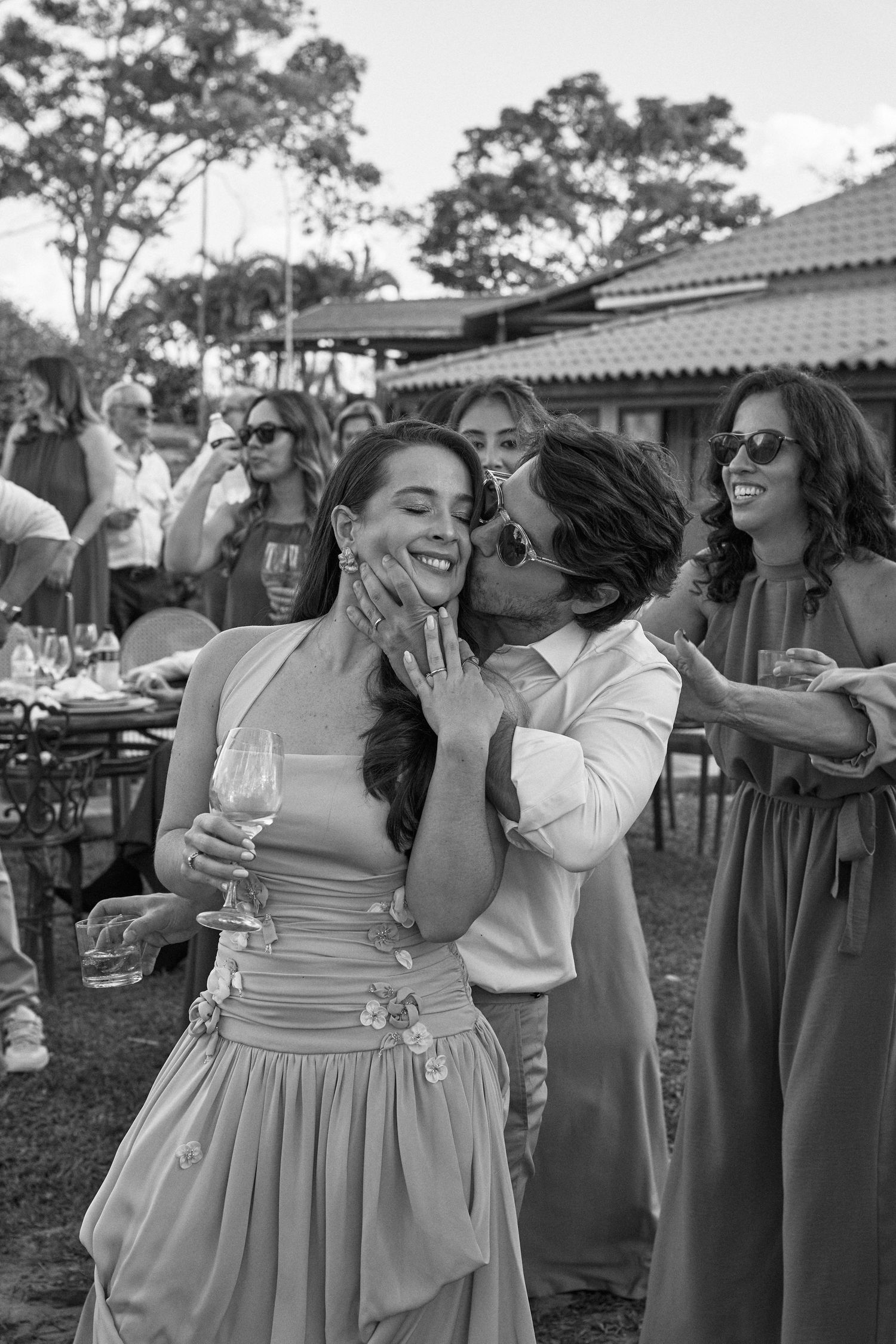 Uma festa de casamento celebra ao ar livre, com amigos se abraçando e rindo enquanto seguram taças de vinho perto de árvores.