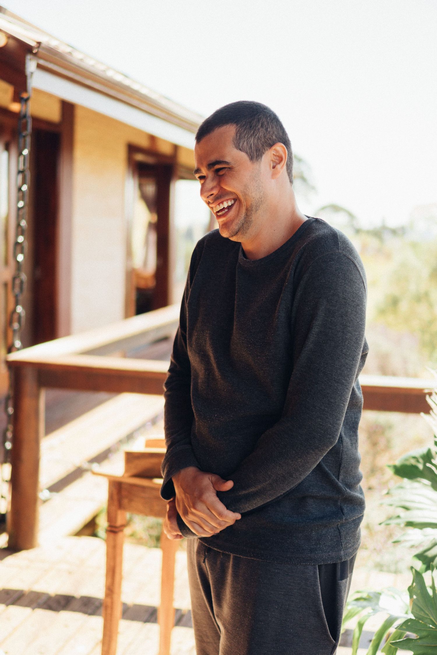 A smiling person wearing a black sweater stands on a sunny outdoor porch.