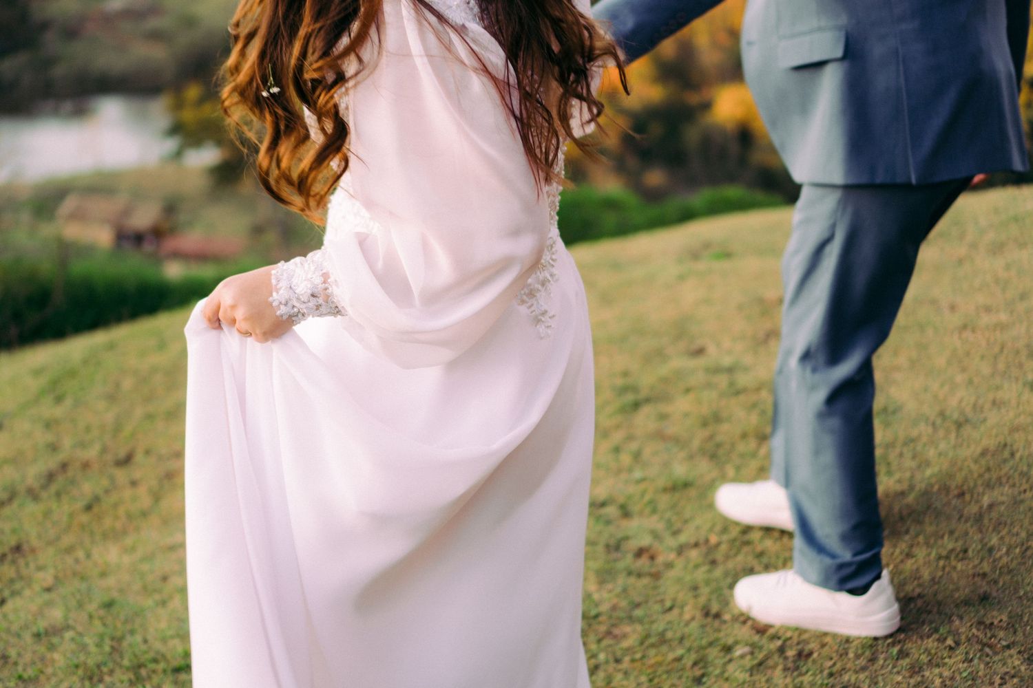Close-up de um vestido de noiva branco esvoaçante com pérolas e detalhes de renda em uma colina gramada.
