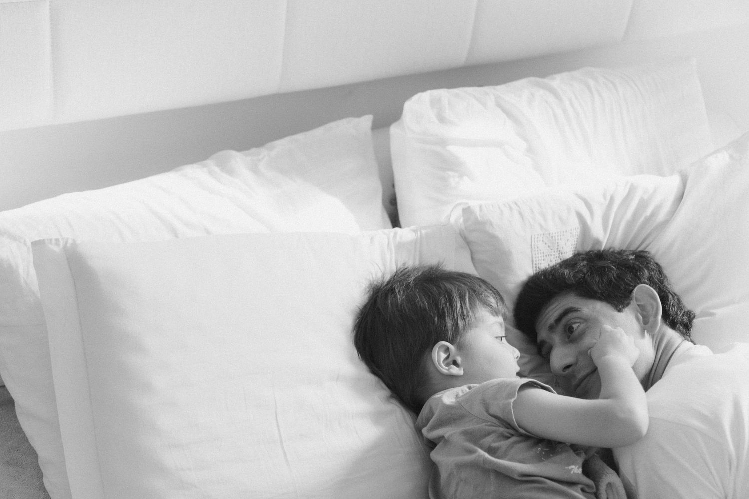 Pai e filho se olham deitados na cama © Clara e Matheus Fotógrafos de Casamentos