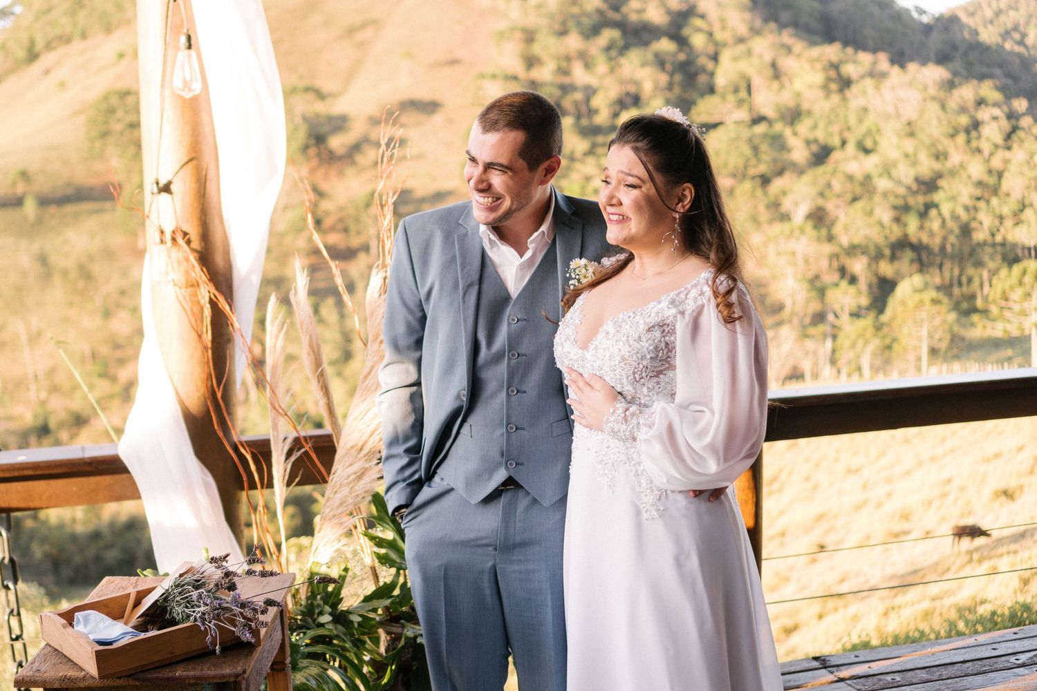 Casal em trajes de casamento elegantes em um deck de madeira com vista panorâmica das montanhas.