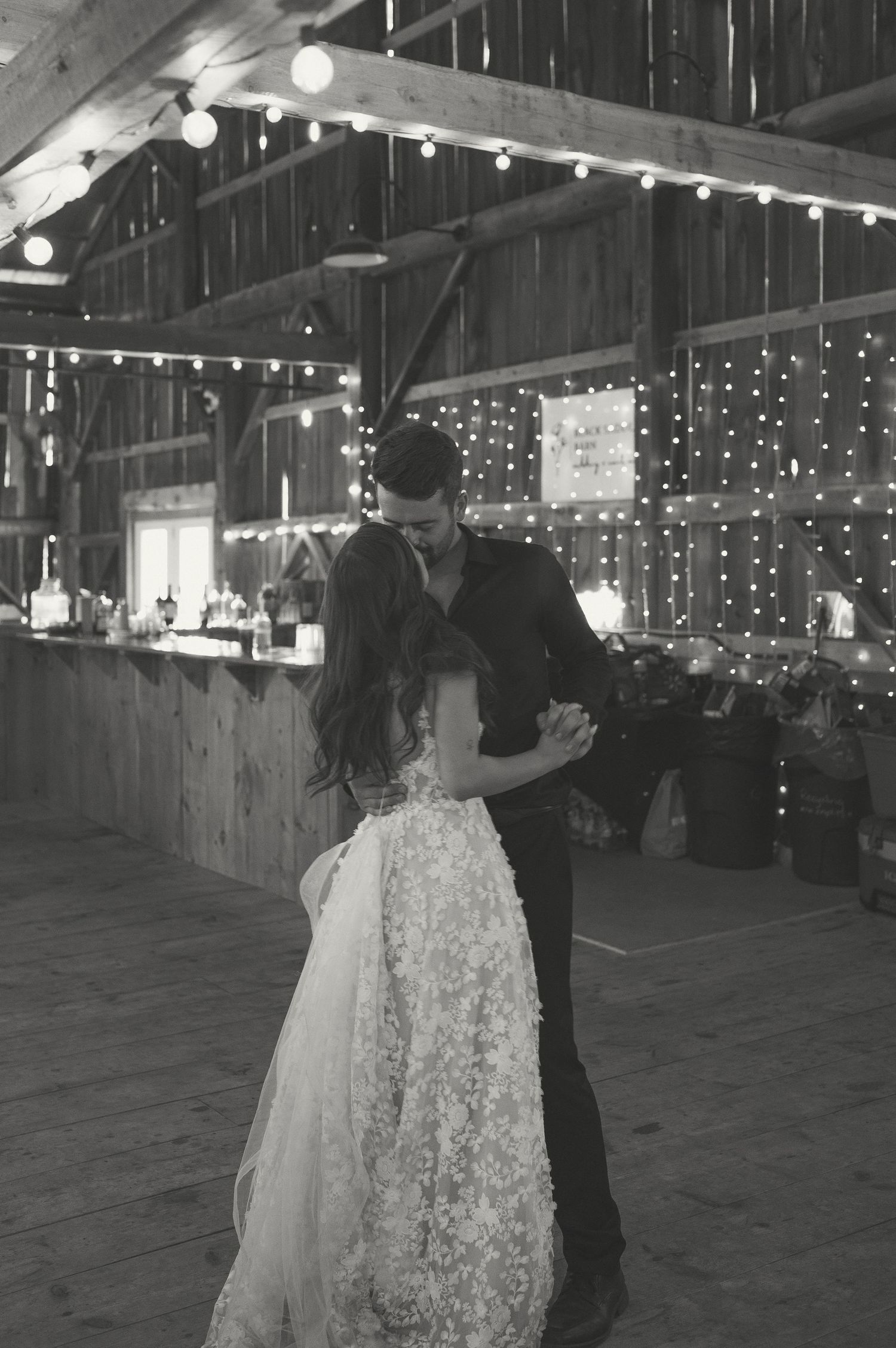 A couple shares a romantic dance in a rustic barn decorated with twinkling string lights for their wedding reception.
