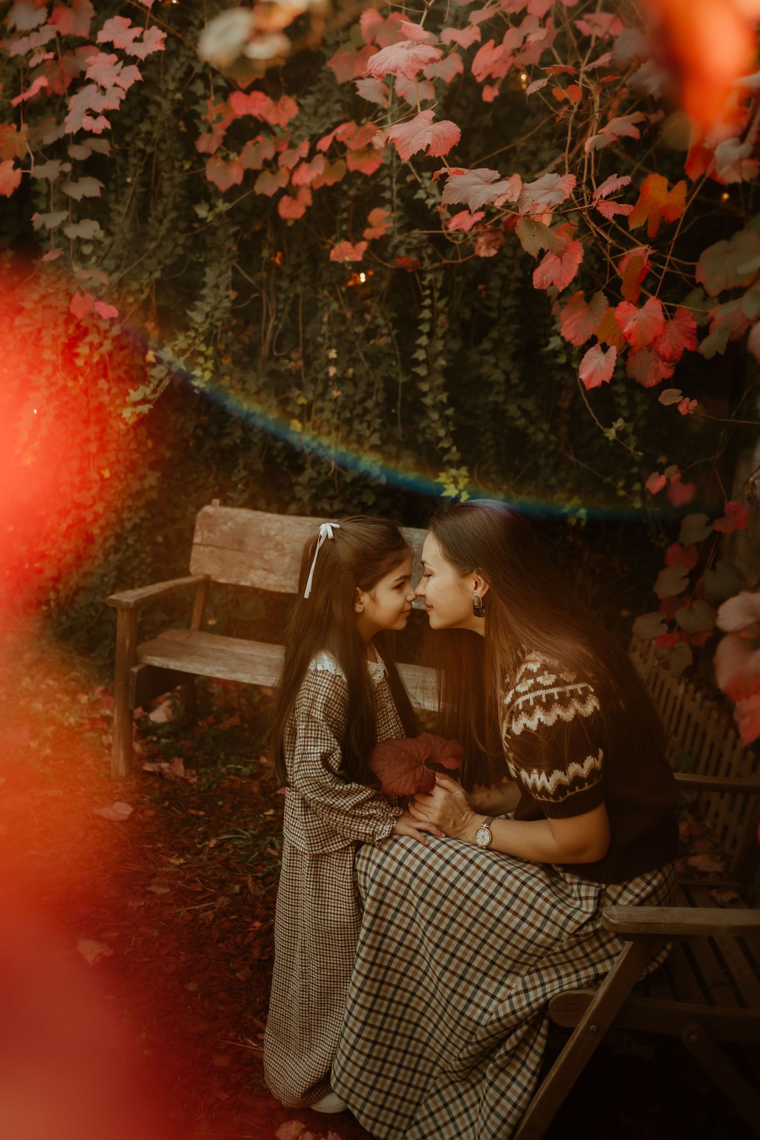 Mother and daughter sharing a tender nose-to-nose moment on a garden bench surrounded by vibrant autumn foliage.