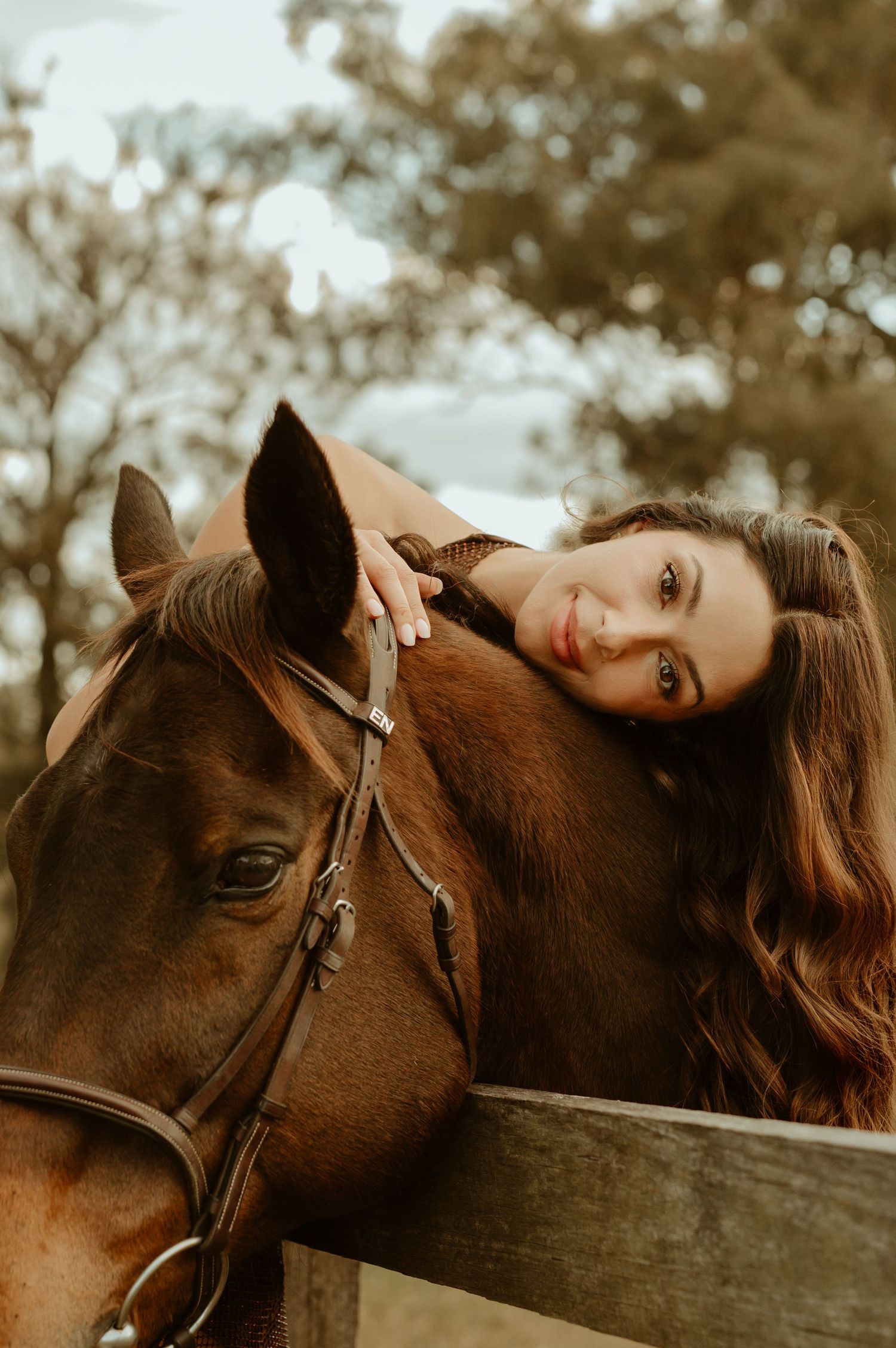 Young woman with long brown hair leans affectionately on a chestnut horse wearing a bridle, outdoors near a fence.