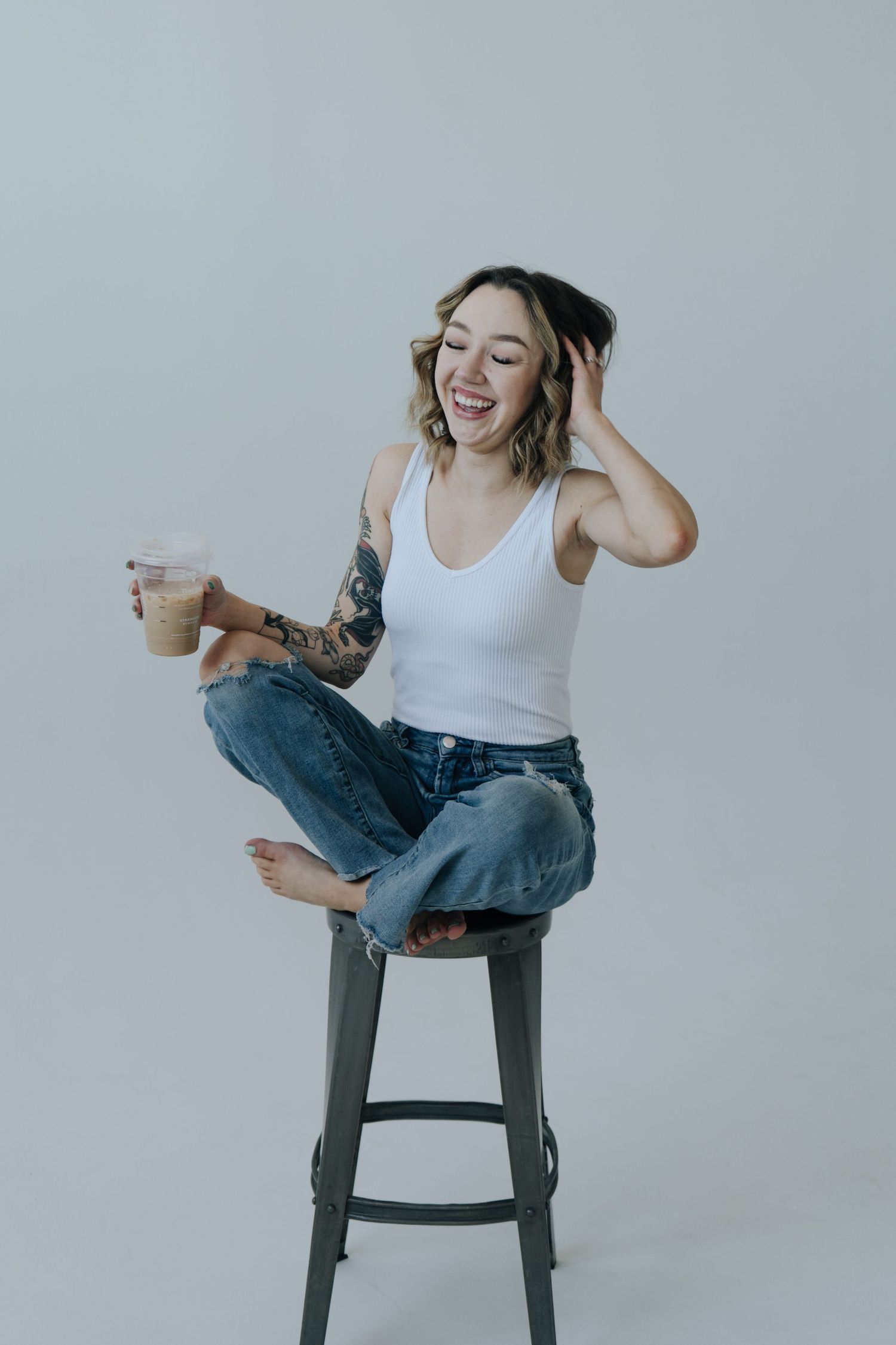 Person in white tank top and jeans sitting casually on dark stool against light background showing playful expressions.