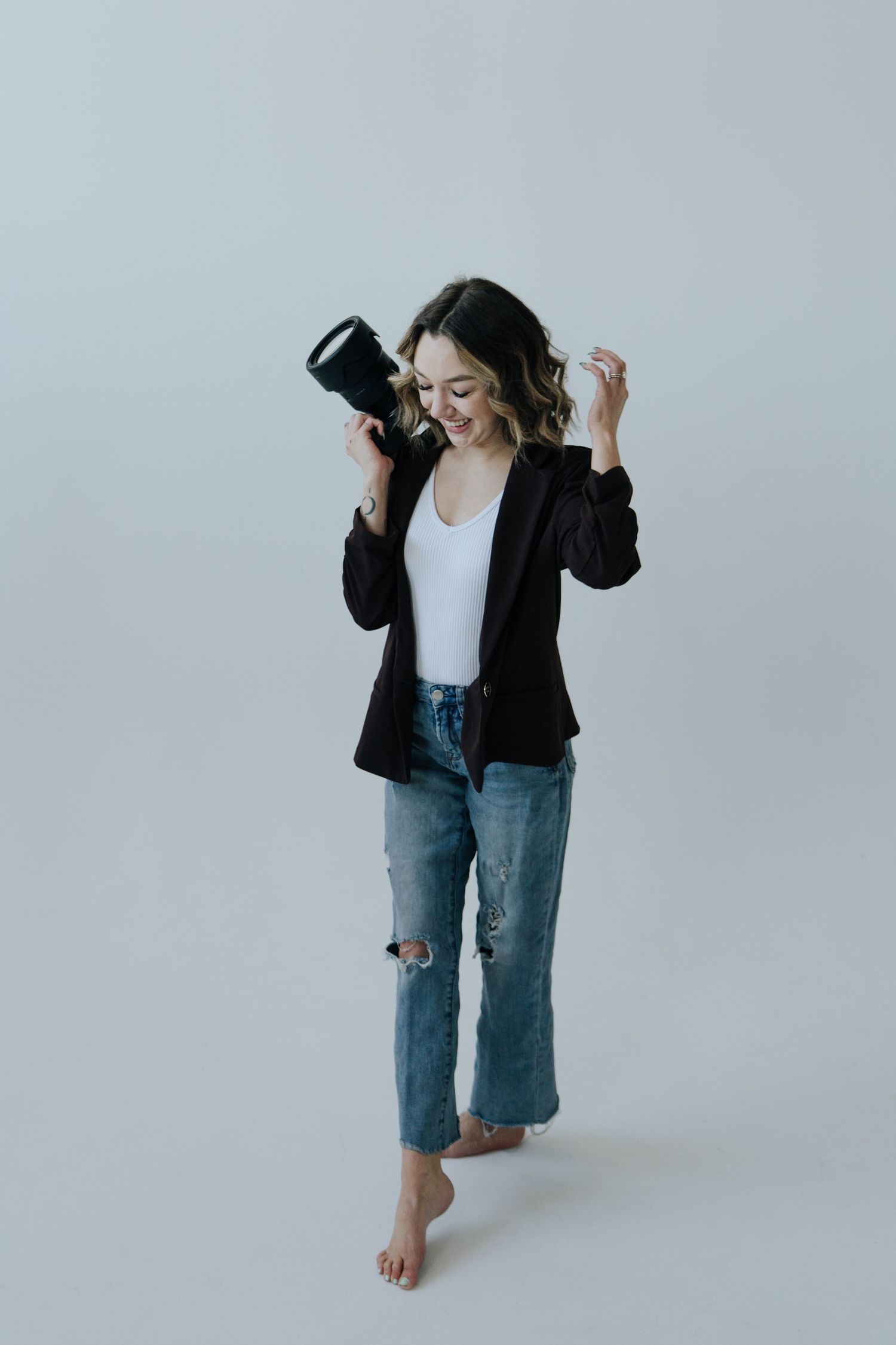 Person wearing casual outfit of black blazer, white top and distressed jeans poses against light gray background.