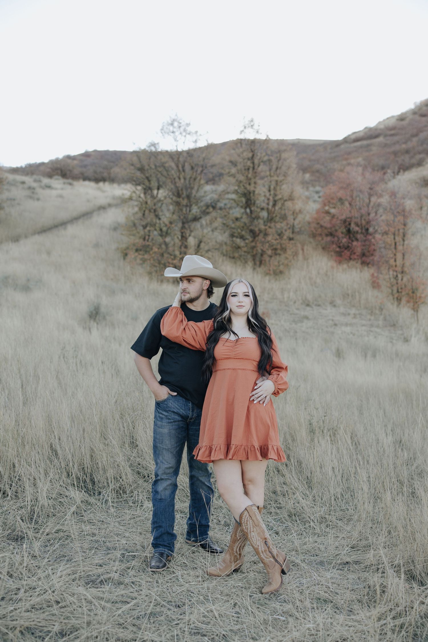 A couple in coordinating orange and black outfits pose together in tall grass on a hillside with fall scenery.