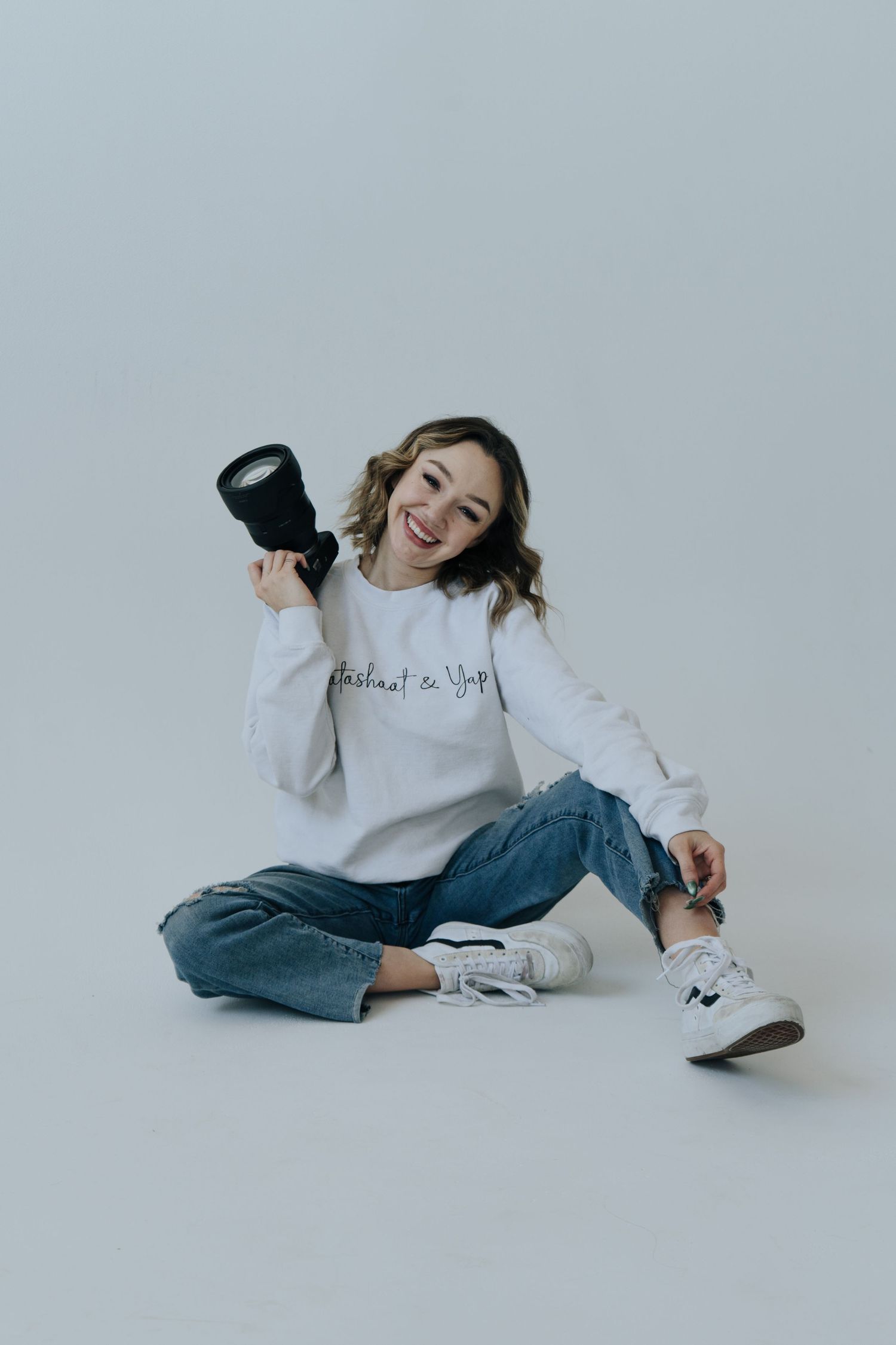 A person in a white sweater and jeans lies playfully on a light gray background in a photo series.
