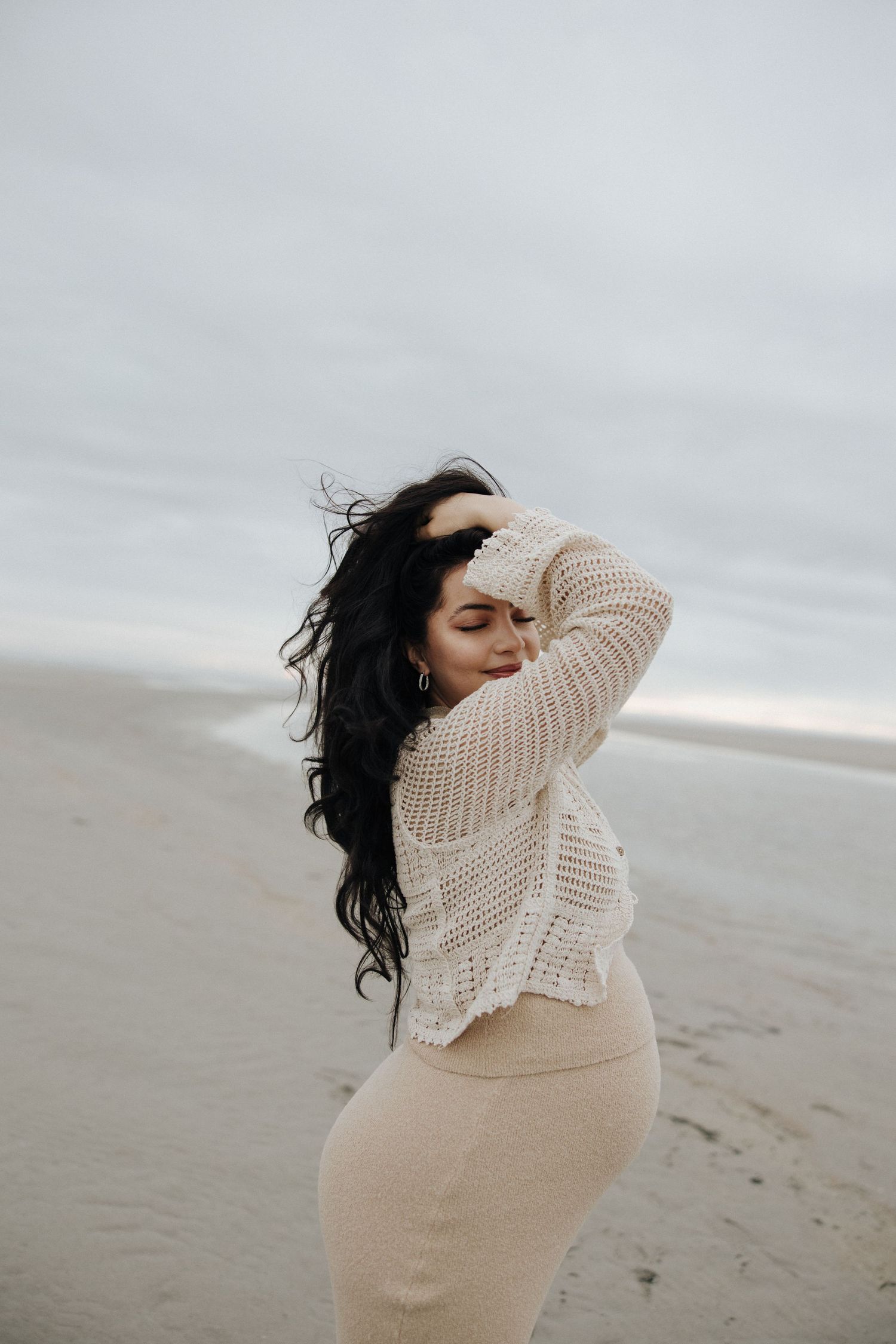 A person in a cream sweater stands on a misty beach with windswept hair creating a dreamy coastal atmosphere.