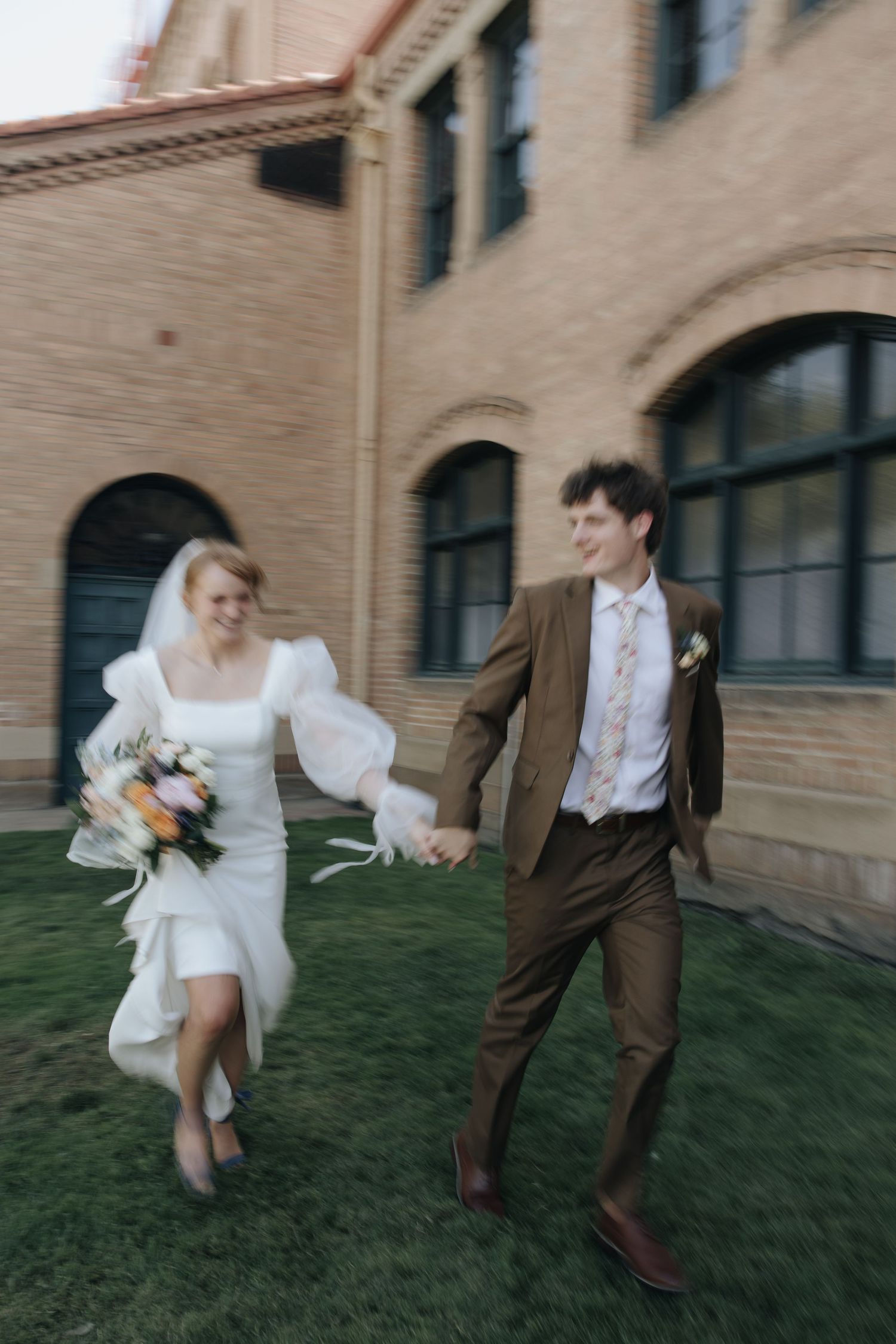 A joyful wedding couple runs across a grassy lawn in front of a historic brick building.
