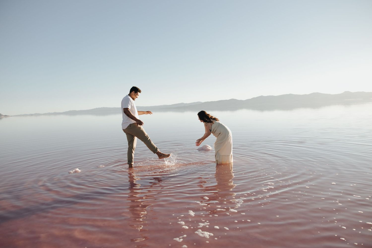Two people splash and play in shallow water at sunset creating rippling reflections in the tranquil surface.