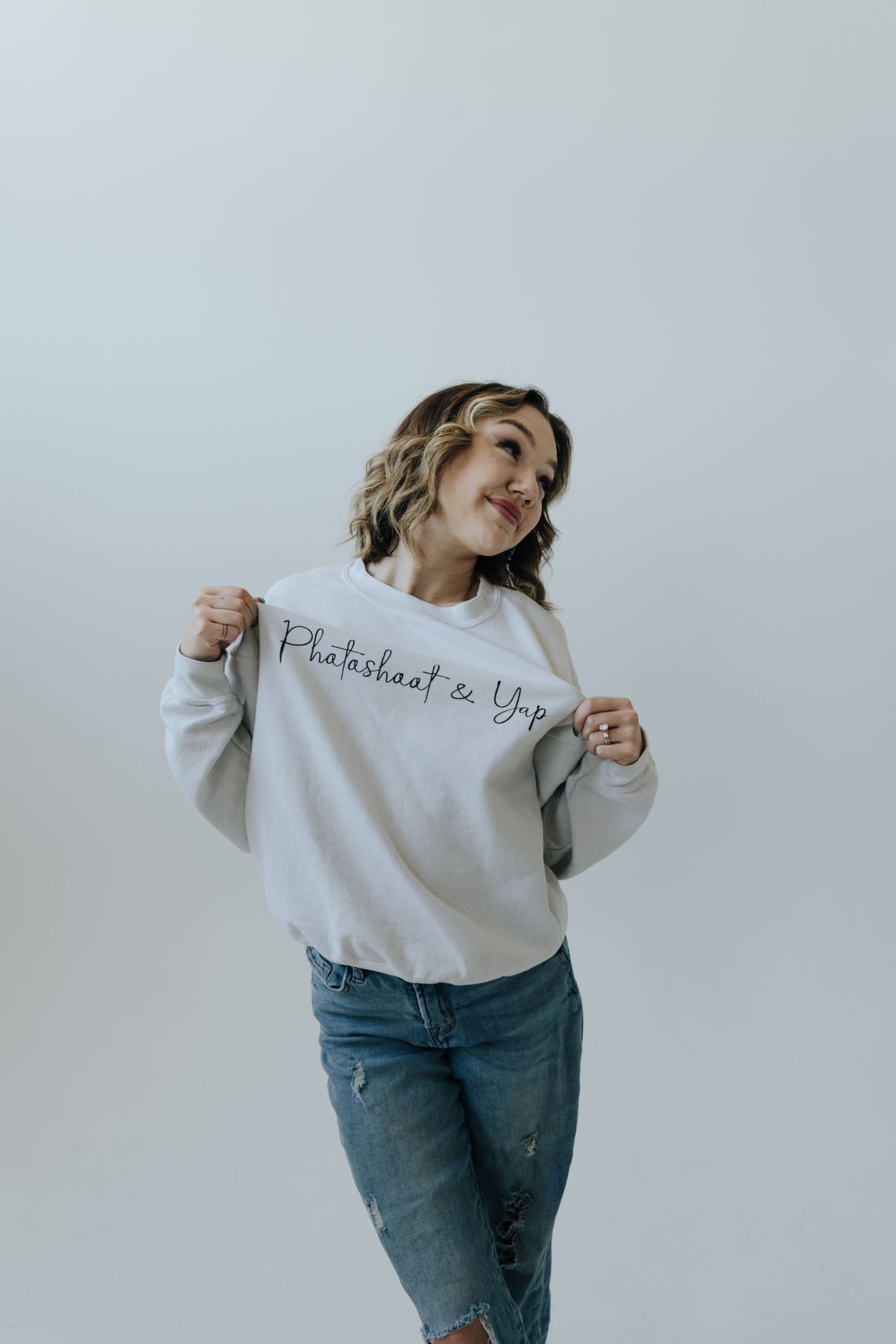 Person wearing a white sweatshirt and distressed jeans poses playfully against a light gray background.