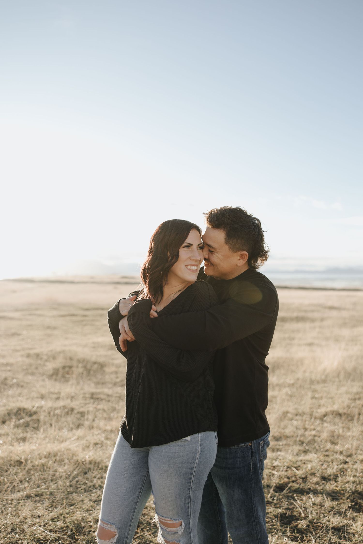 A couple embraces and laughs together in a golden field during sunset, wearing casual denim and black attire.