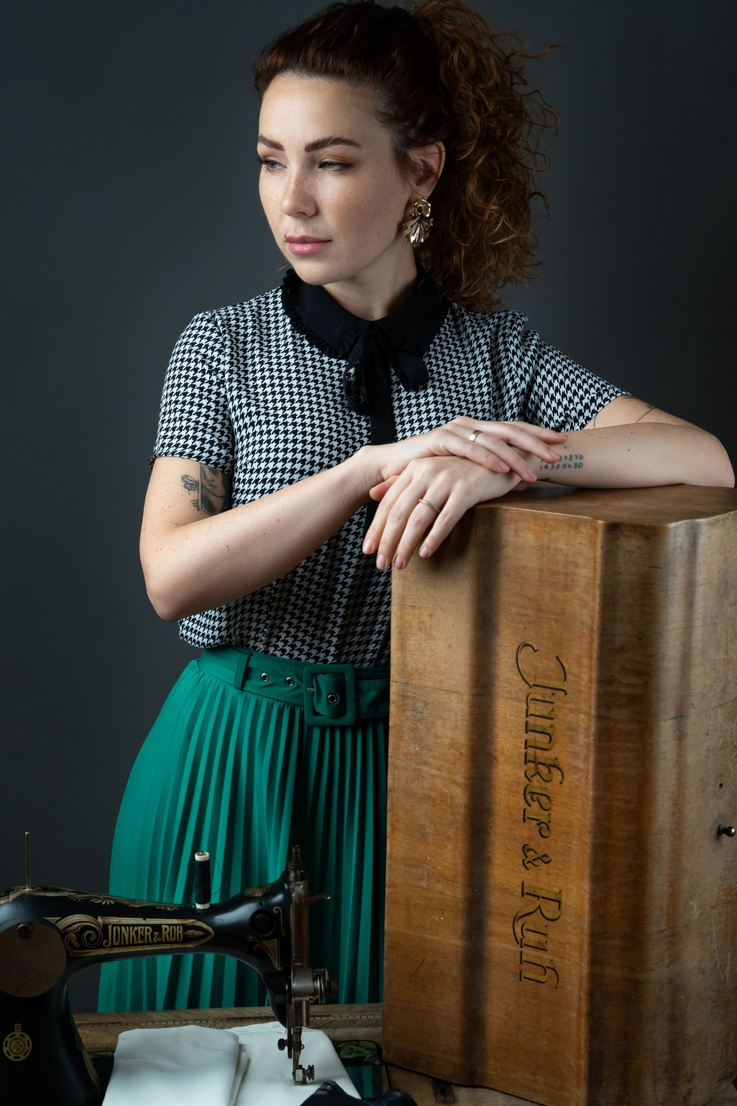 Woman in houndstooth top and green skirt leans on vintage Singer sewing machine cabinet in studio portrait.