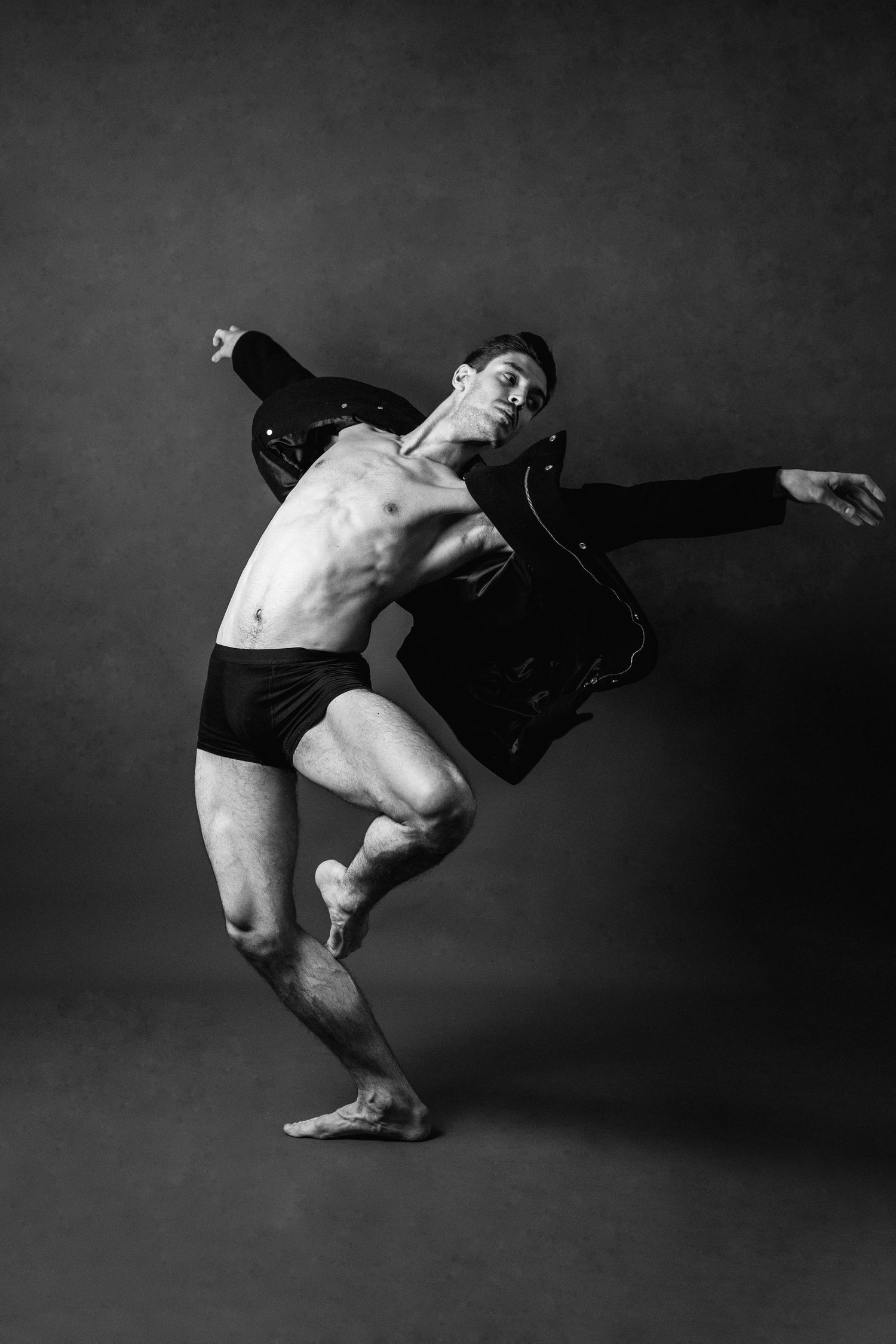 Black and white photo of a male dancer mid-pose, wearing black shorts and jacket, showcasing athletic flexibility.