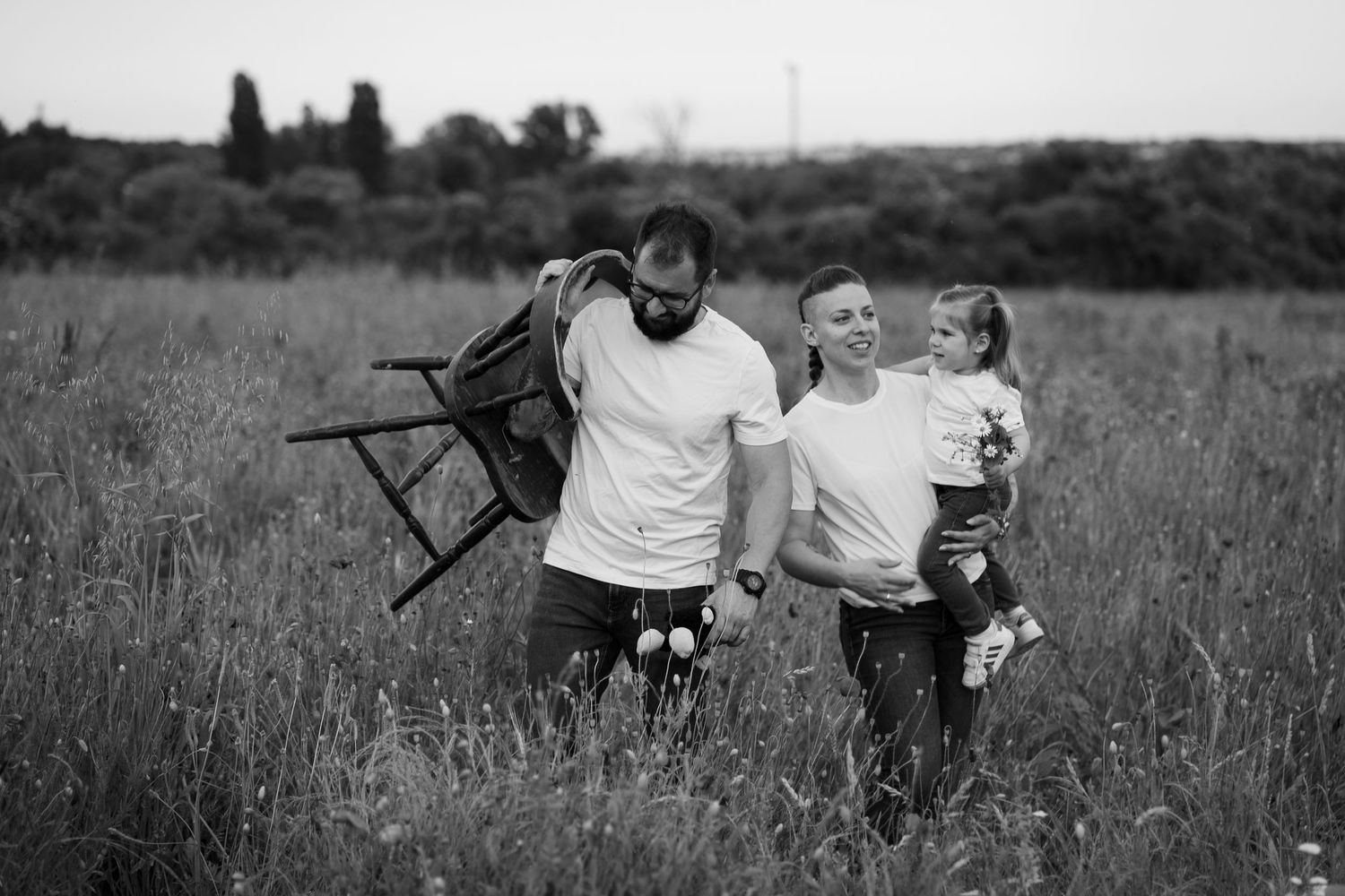 Black and white photo of a family walking through a field, man carrying a chair, woman holding a child with flowers.