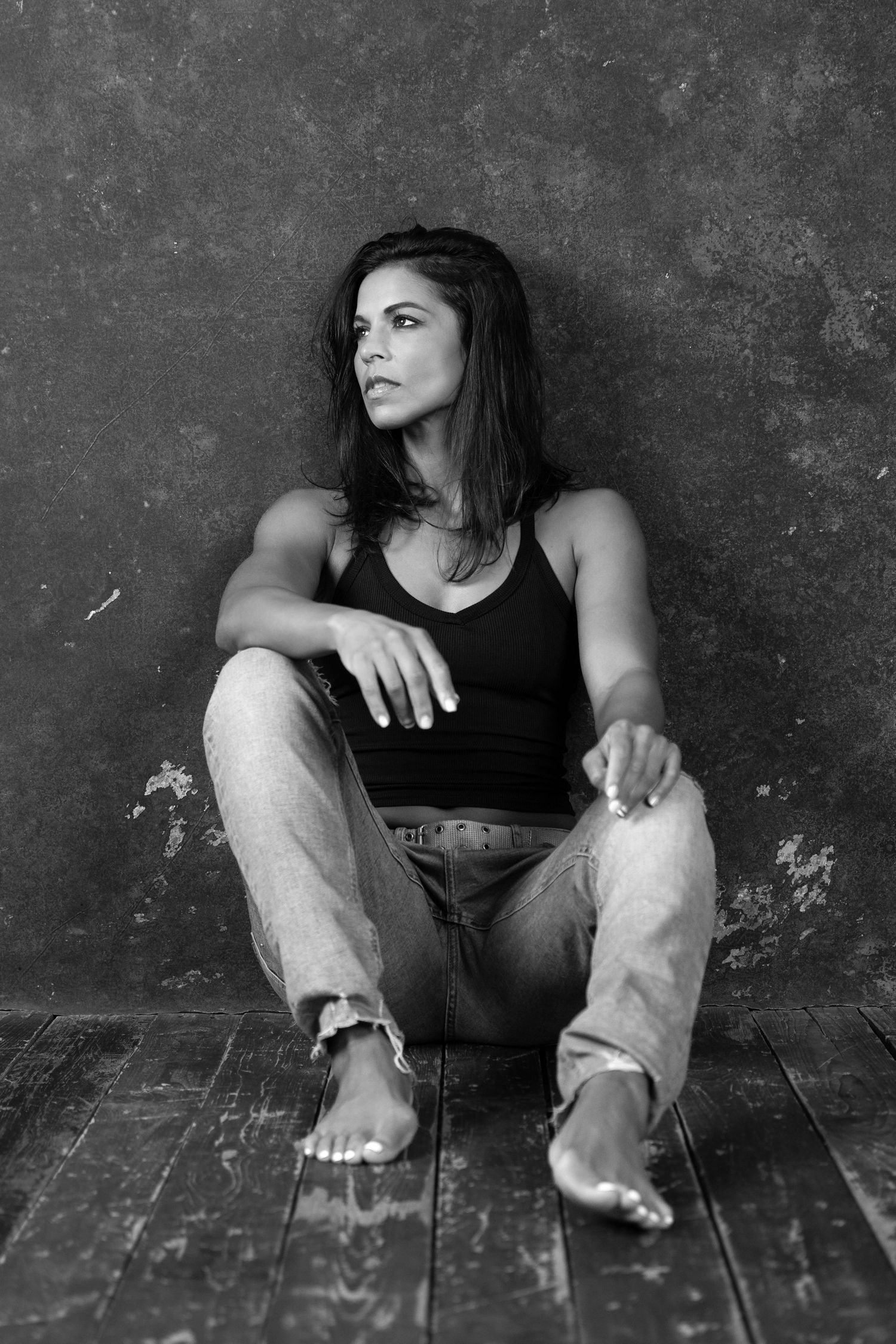 Black and white portrait of a woman sitting on wooden floor in jeans and tank top against grunge wall.