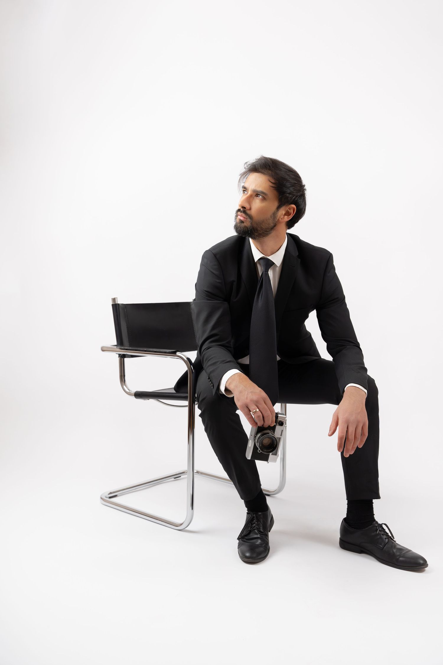 A bearded businessman in a black suit sits on a modern chair, holding sunglasses, gazing thoughtfully on white background.