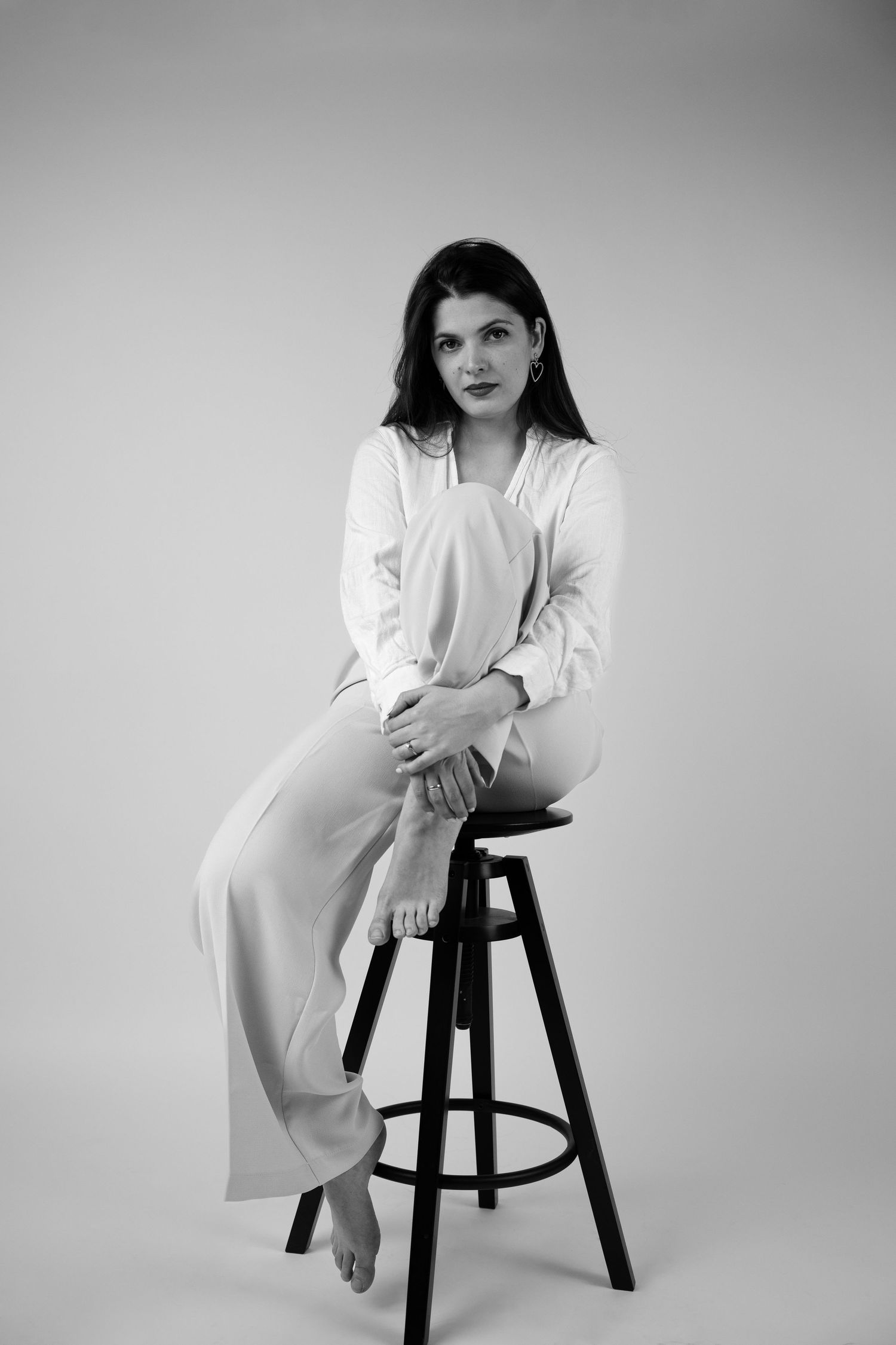 Black and white portrait of a young brunette woman sitting on a bar stool in a white outfit, barefoot.