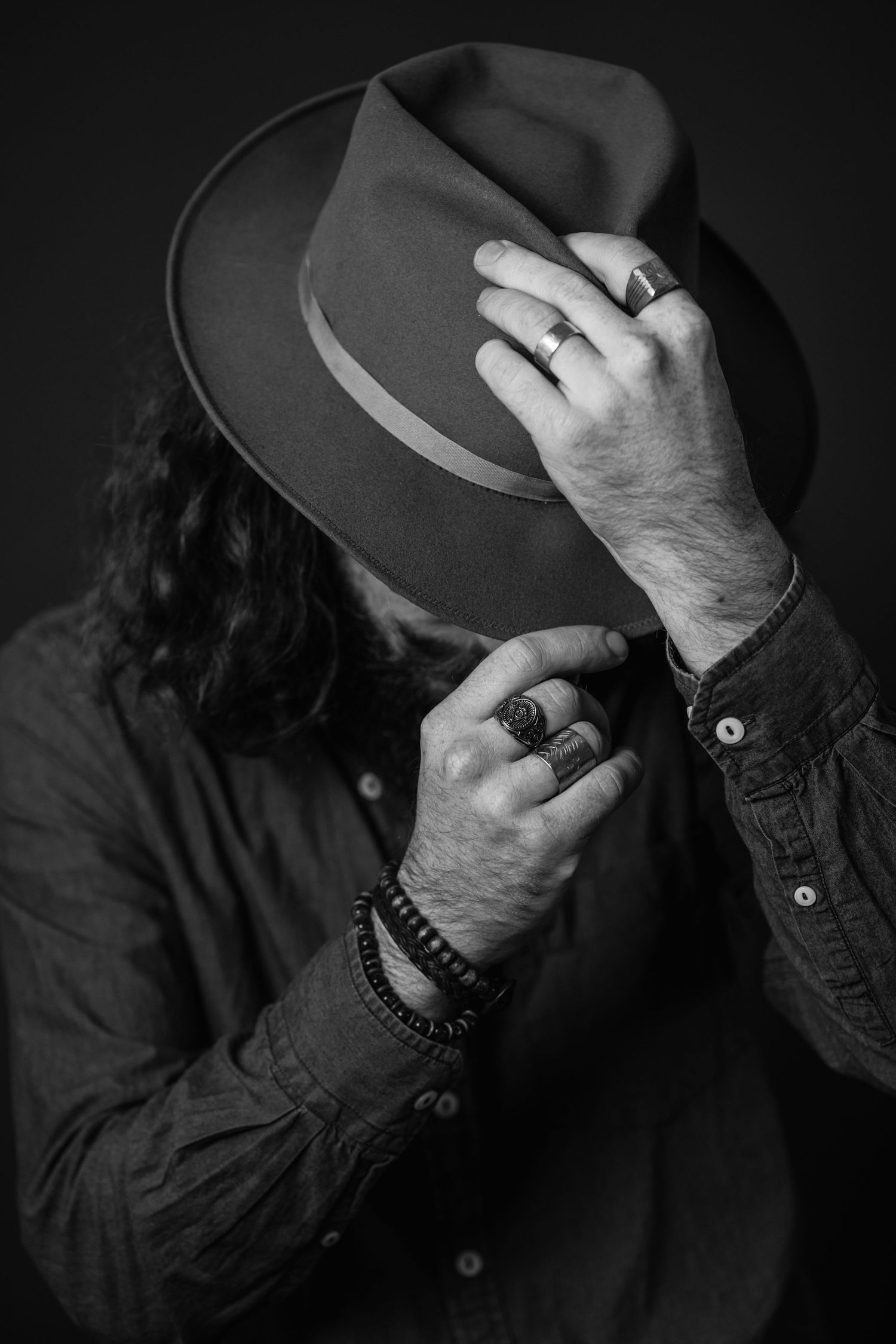Black and white portrait of a long-haired man hiding his face behind a wide-brim hat, wearing rings and a beaded bracelet.