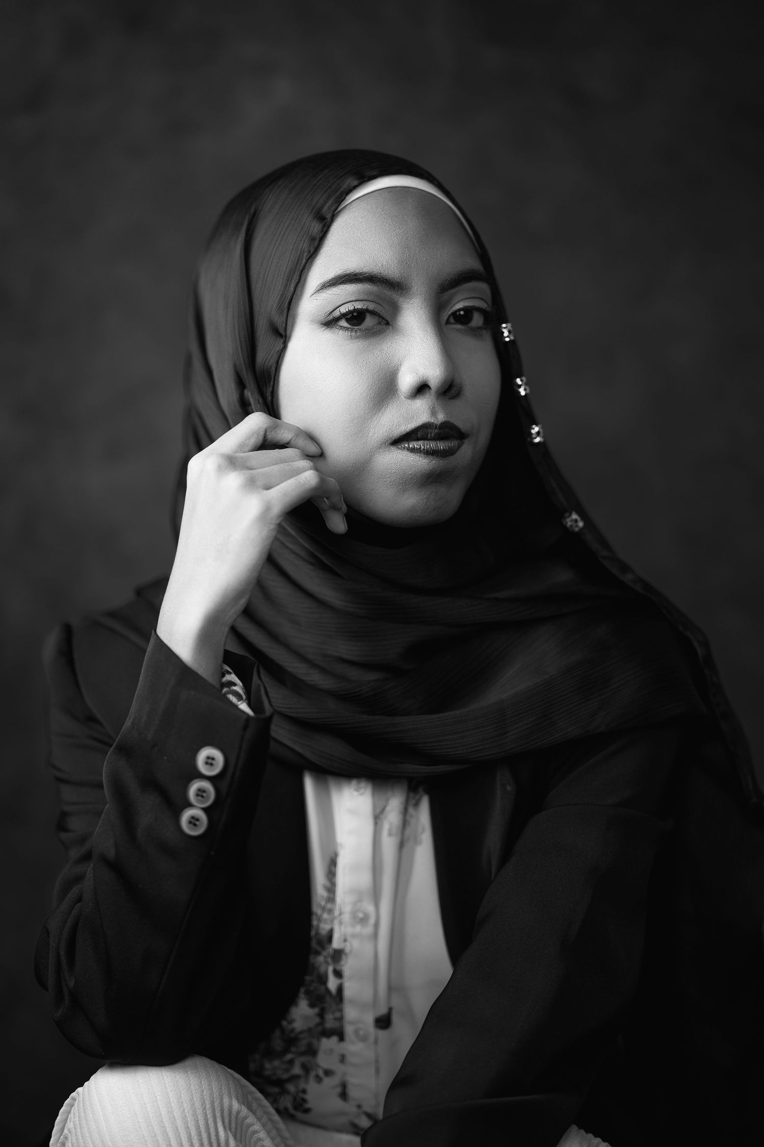 Black and white portrait of a young woman wearing a hijab and blazer, posing gracefully against a dark background.