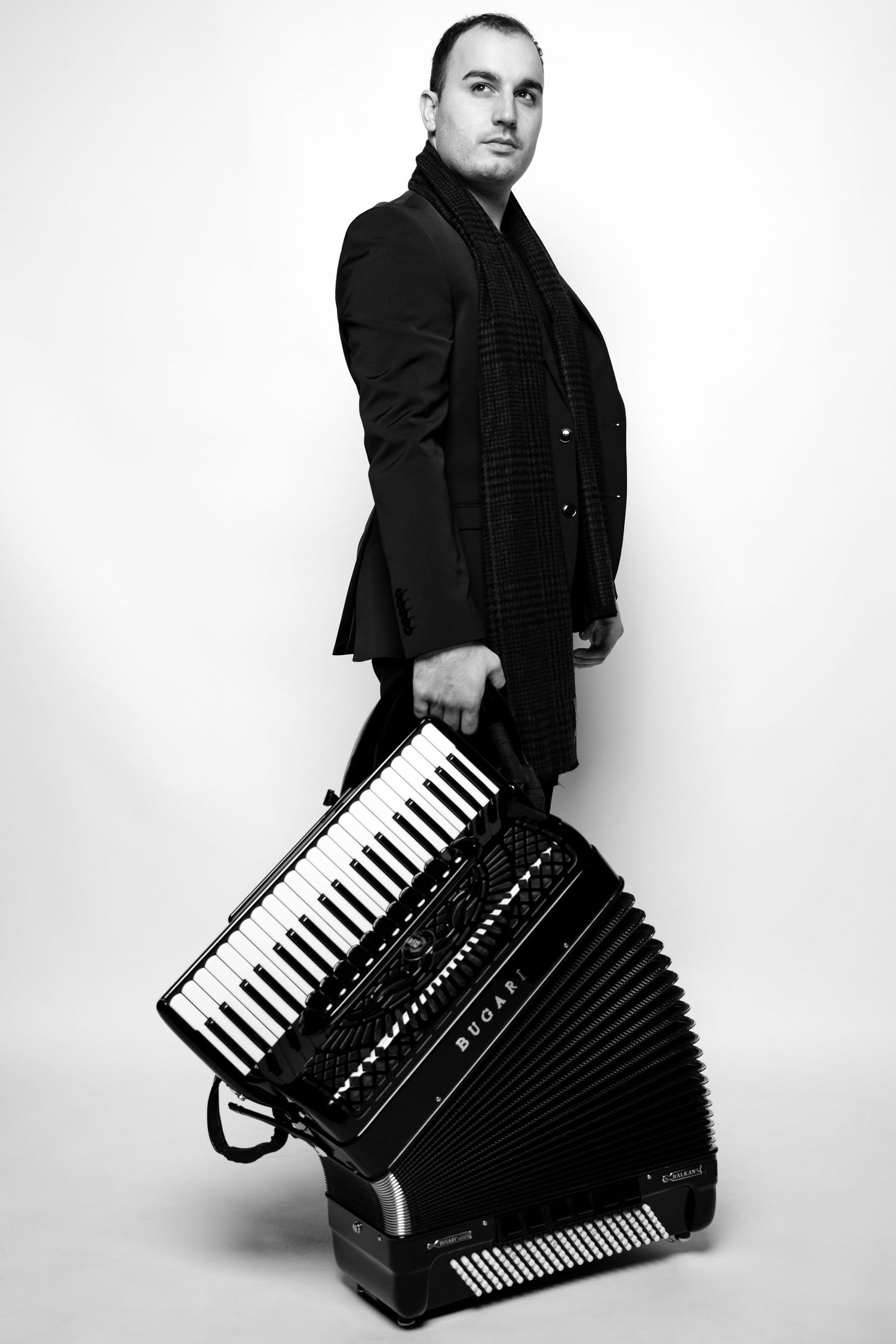 Black and white photo of a man in a dark suit holding an accordion instrument while posing against a white background.