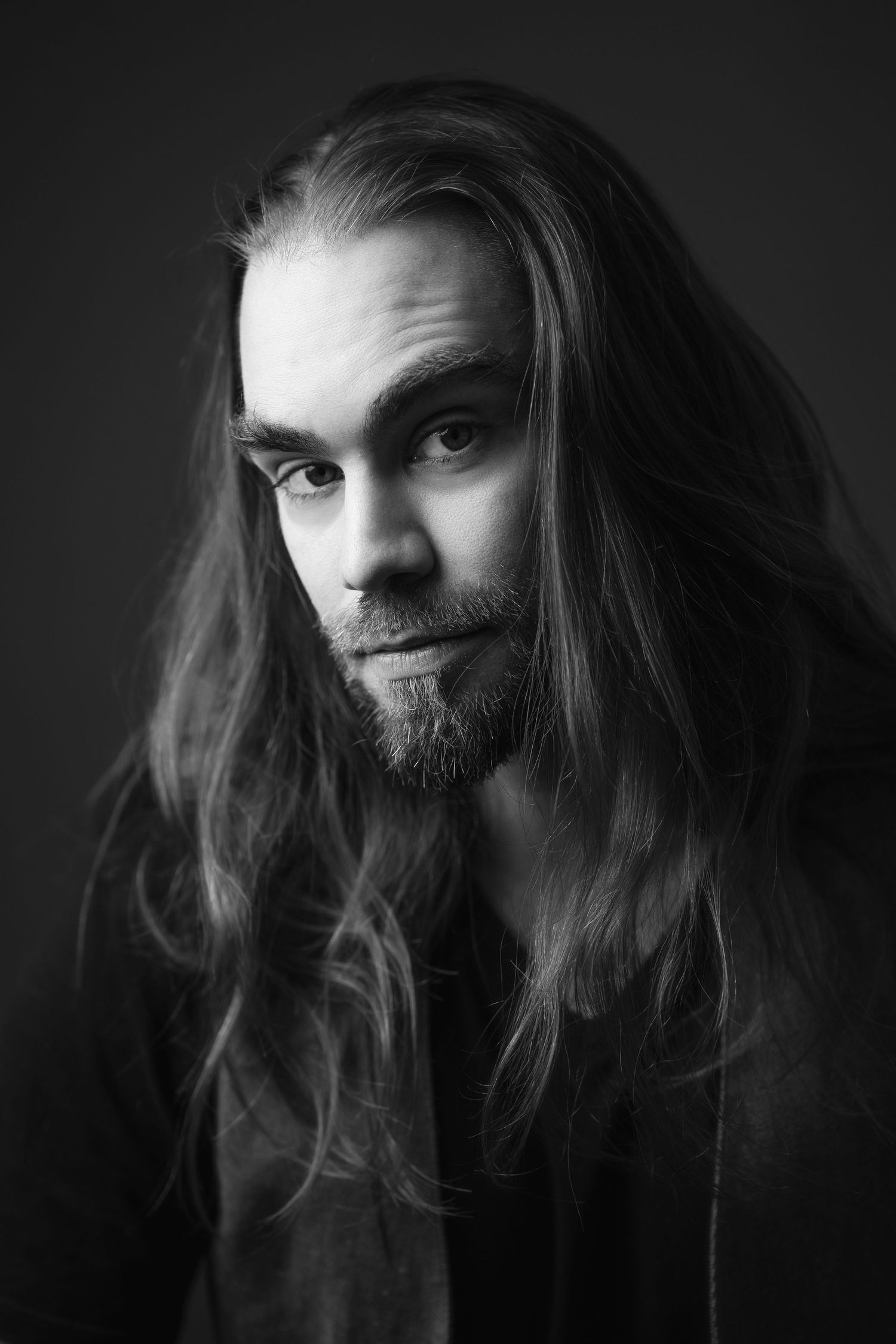 Black and white portrait of a young man with long flowing hair and beard against dark background.