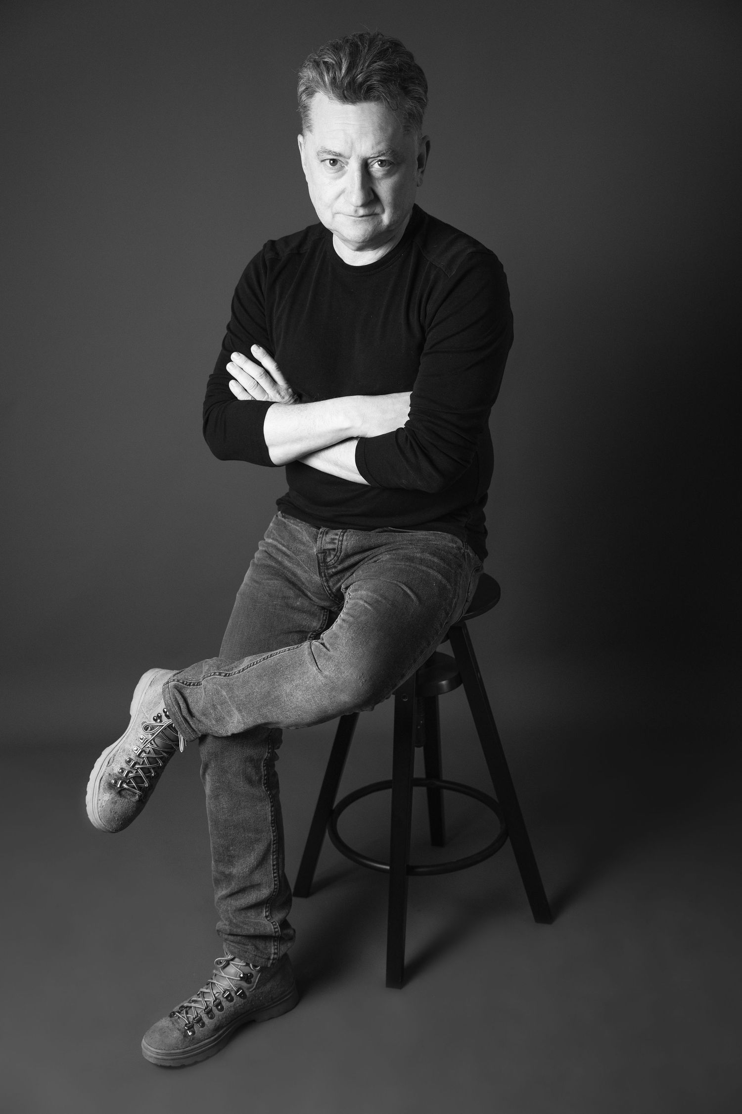 Black and white portrait of a middle-aged man sitting on a stool with arms crossed, wearing jeans and a dark sweater.