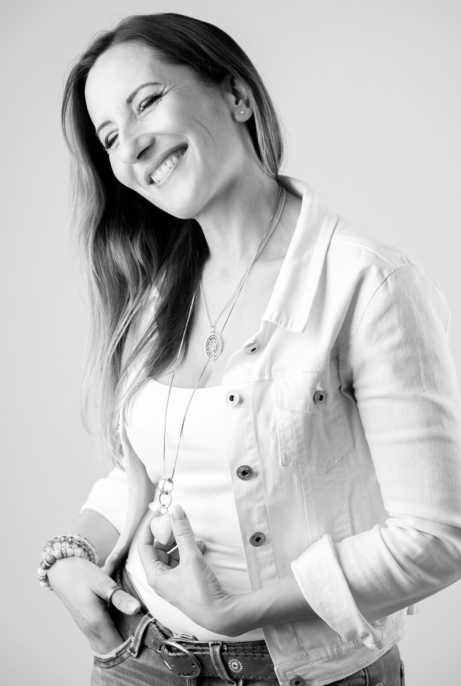 Black and white portrait of a smiling woman in a white jacket and jeans, wearing layered necklaces and a bracelet.